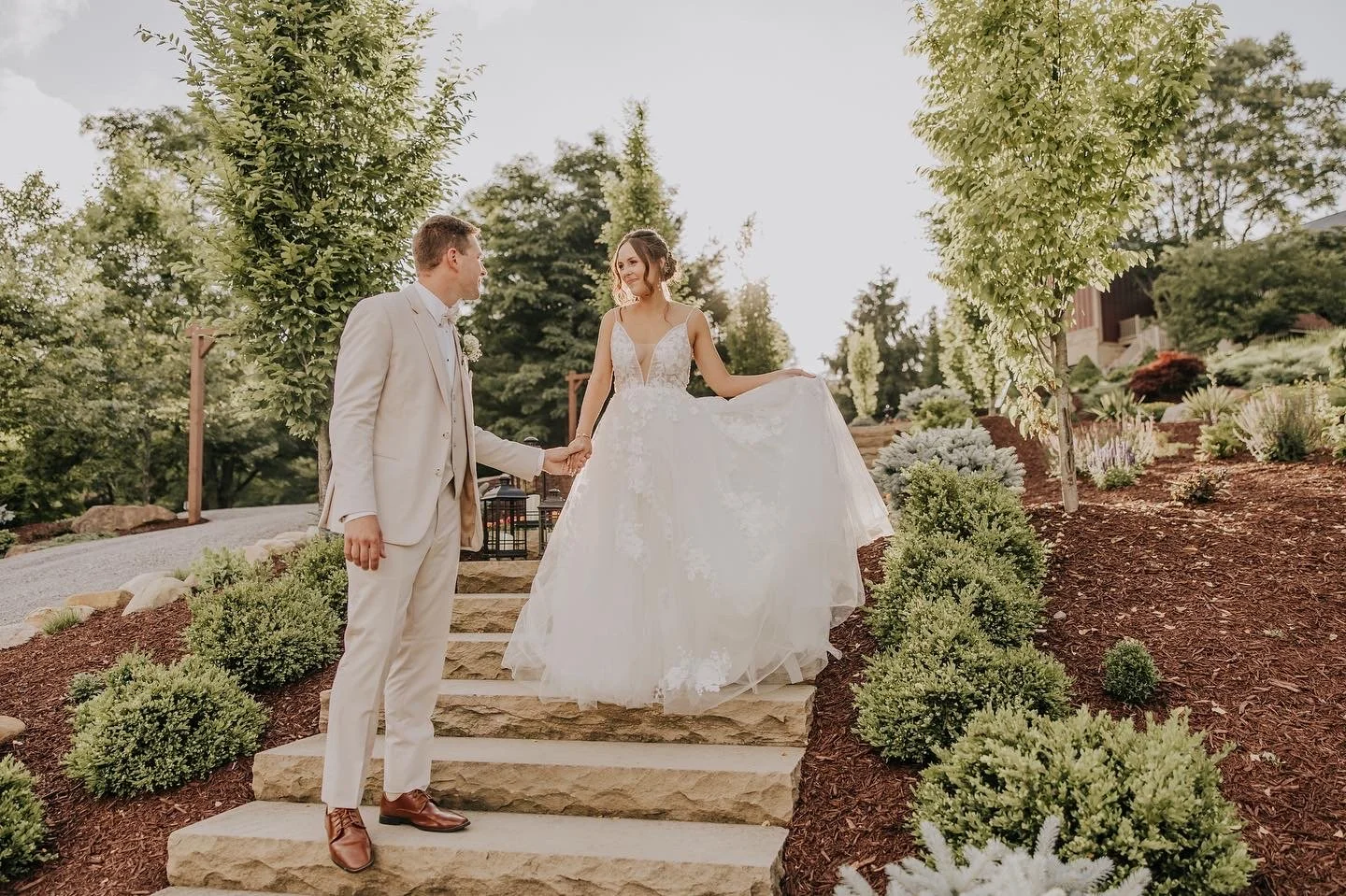 A bride and groom holding hands on stone steps in a lush outdoor garden, with sunlight filtering through trees.