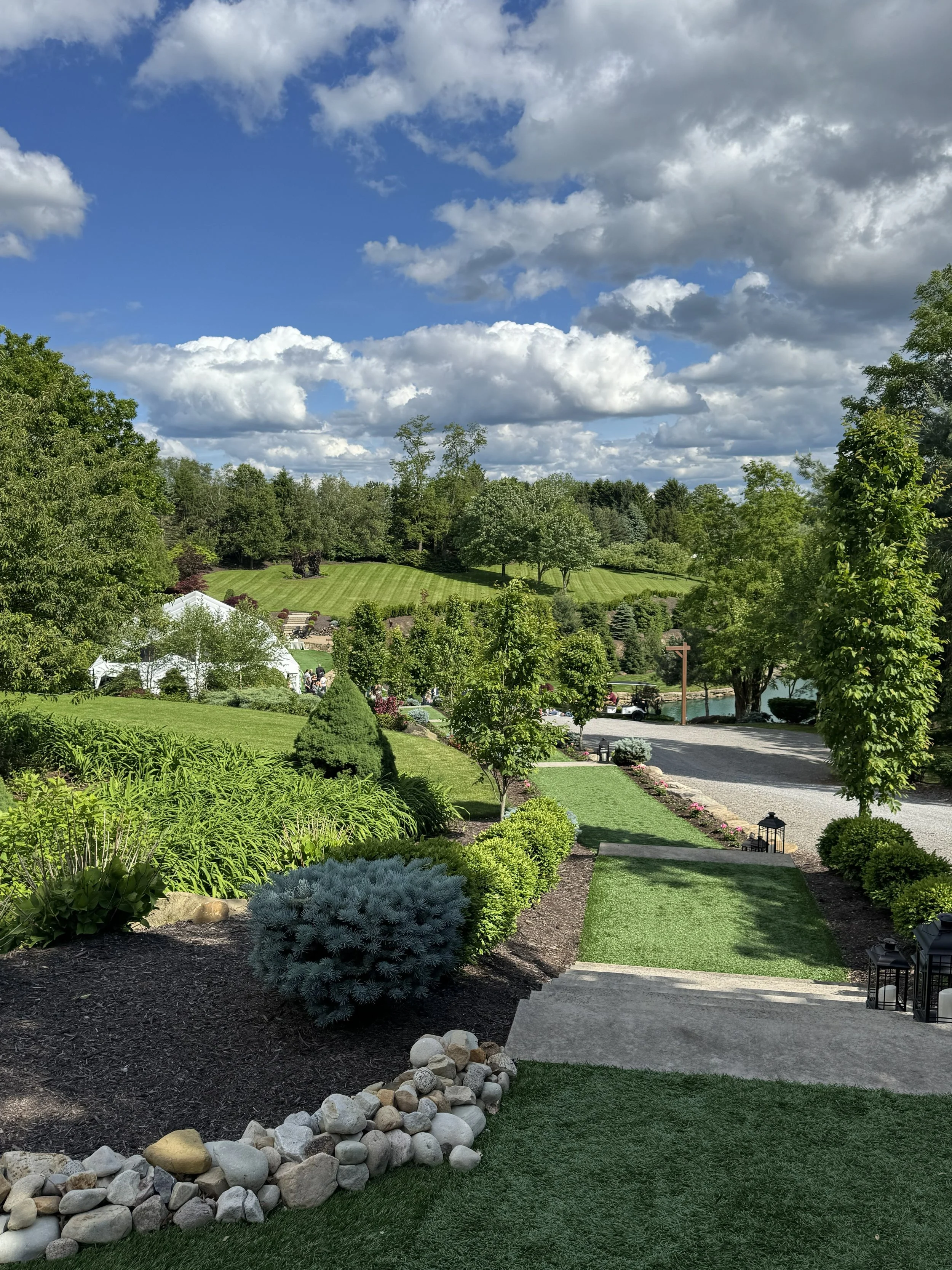 Beautiful landscaped garden with a lawn, trees, shrubs, a gravel pathway, decorative rocks, and a distant view of rolling hills under a partly cloudy sky.
