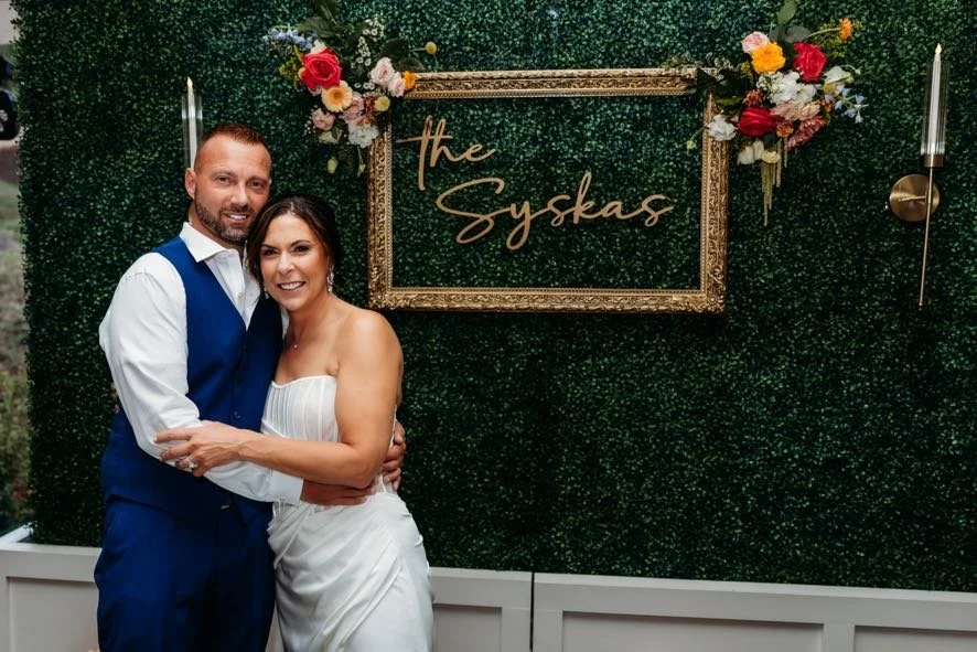 A couple in wedding attire smiling and hugging in front of a green floral backdrop with a gold frame sign that reads 'the Syskas' and decorated with flowers and candles.