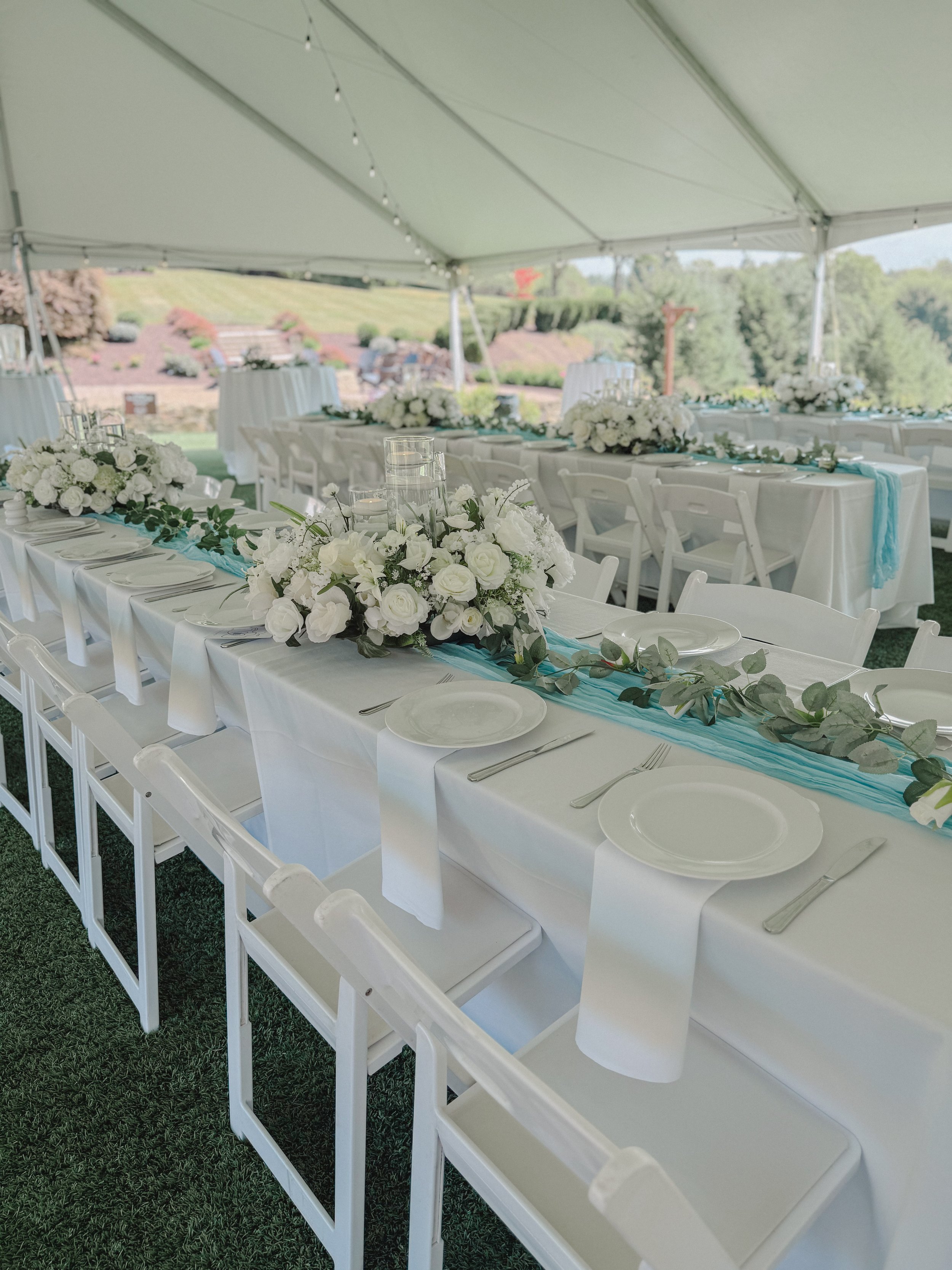 Elegant outdoor wedding reception setup under a tent with white tablecloths, floral centerpieces, and white chairs.
