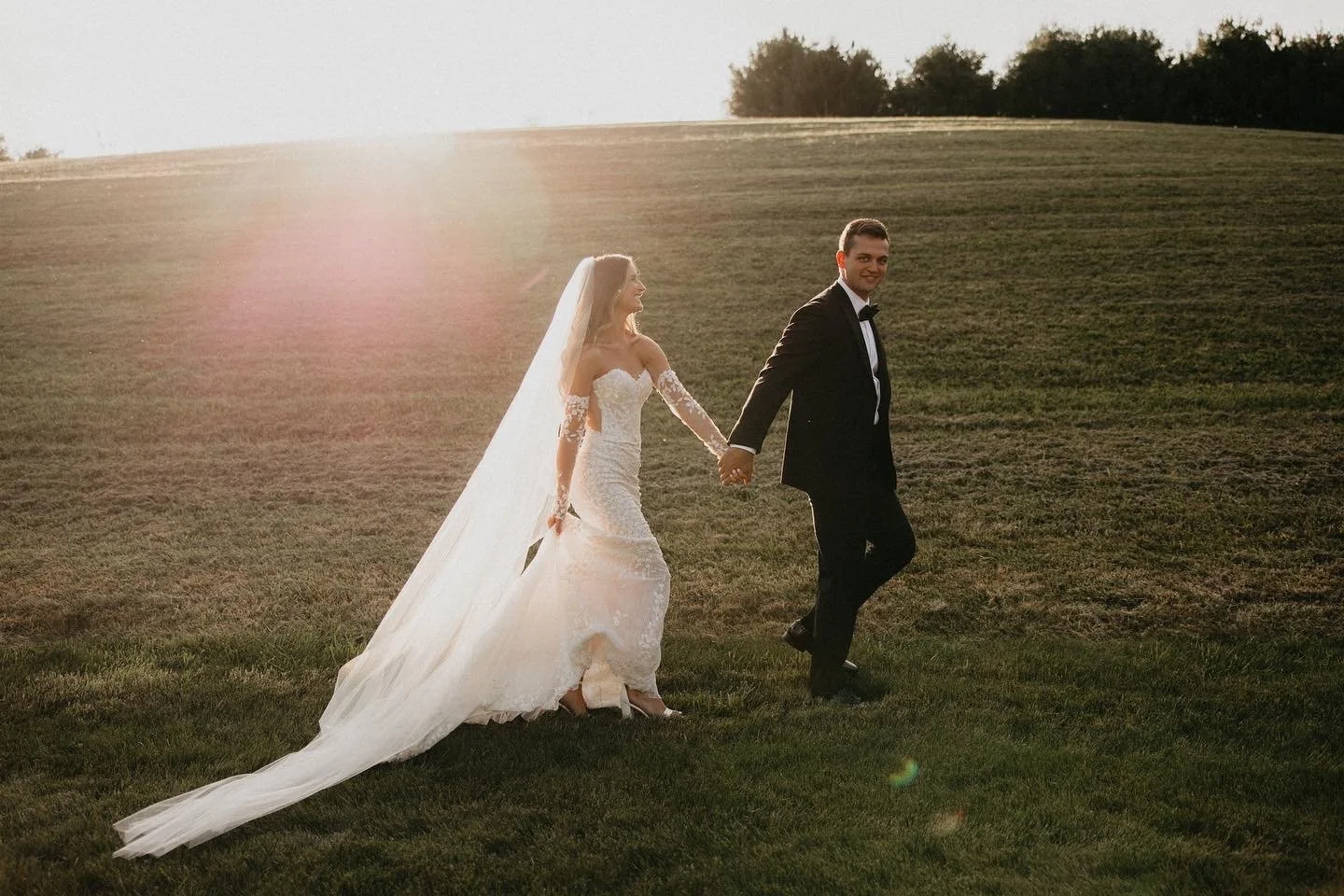 A bride and groom holding hands, walking on a grassy field at sunset, with the bride wearing a white lace wedding dress and veil, and the groom in a black tuxedo.