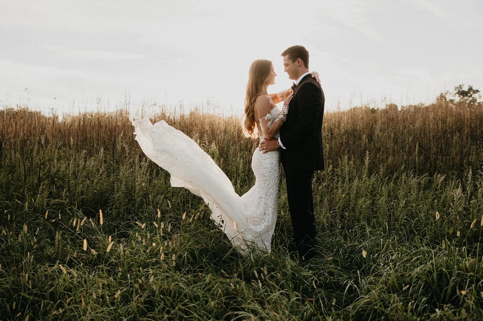 A newlywed couple stands close in a field of tall grass and wildflowers, with the bride in a white wedding gown and the groom in a black tuxedo, during sunset.