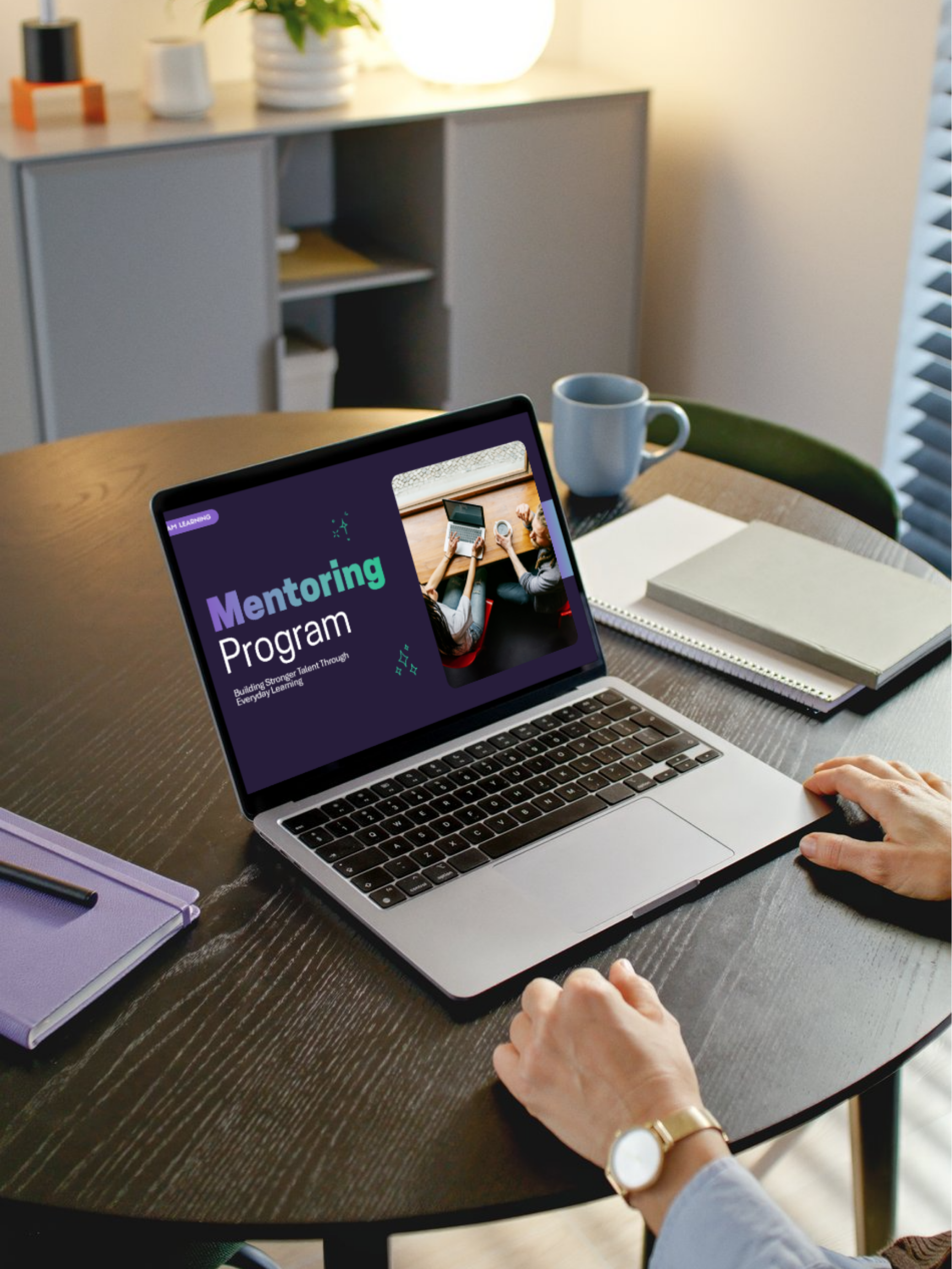 Laptop on a wooden table displaying a mentoring program presentation, with notebooks, a coffee mug, and a person's hand visible.