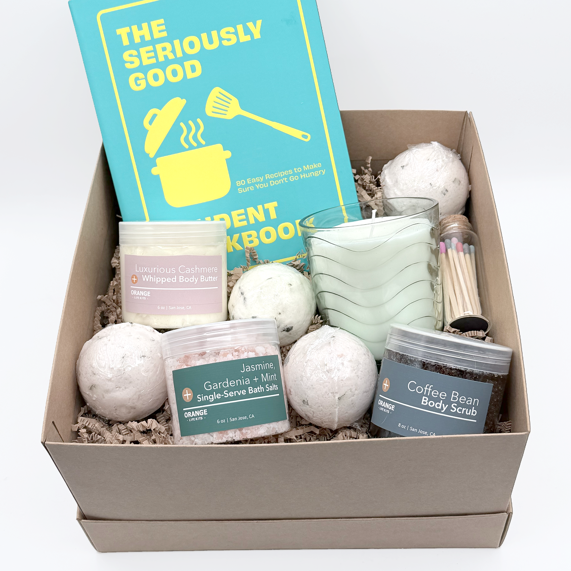 A cardboard gift box filled with self-care items including a blue book titled 'The Seriously Good I Cookbook', a white jar labeled 'Luxurious Cashmere + Whipped Body Butter', a round bath bomb, a clear glass measuring cup with a striped pattern, a small container of jasmine and gardenia bath salts, and a tube of coffee bean body scrub.