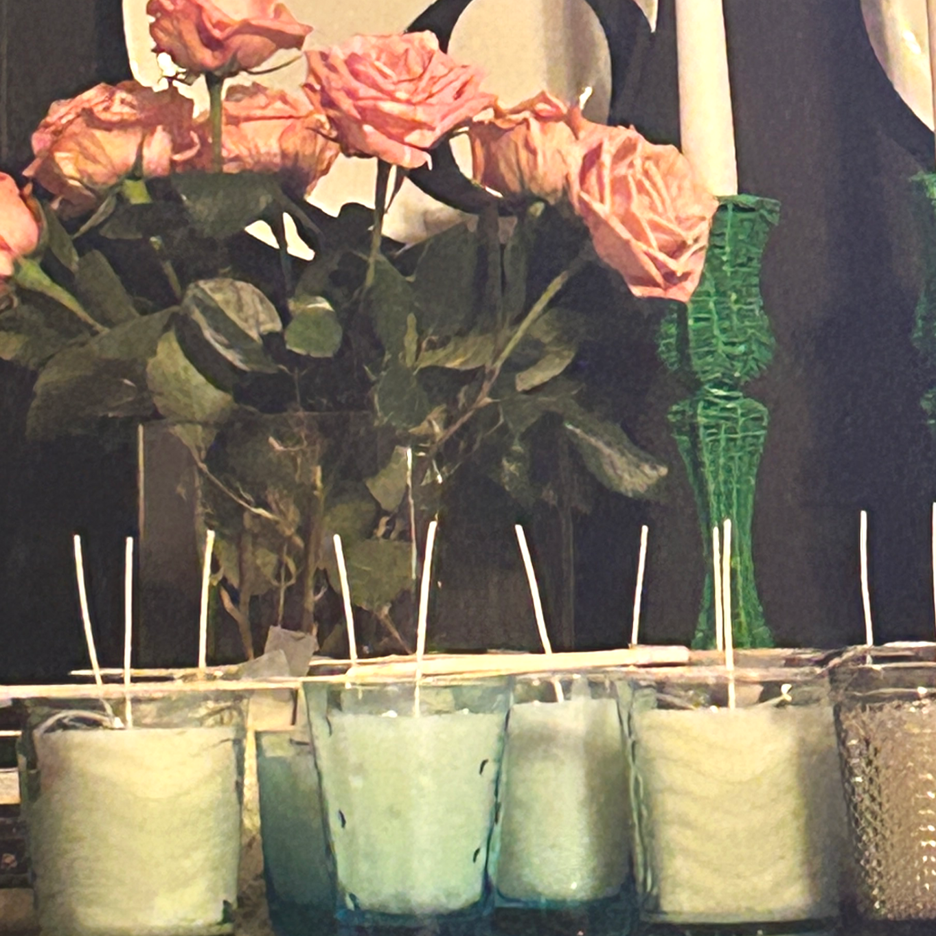 A row of potted plants with pink roses and wilted flowers, with a green glass vase and wooden sticks in the background.