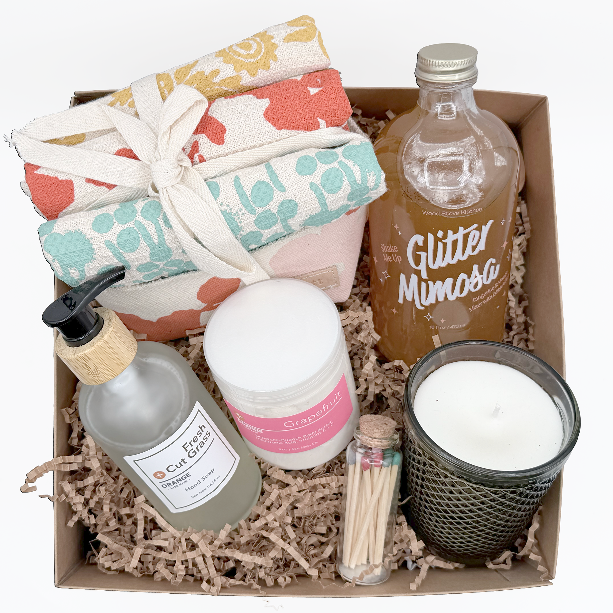 A gift box containing various self-care items including a wrapped towel, a natural brush, bottles of hand soap and moisturizer, a small jar, a glass bottle of drink labeled 'Grapefruit Mimosas,' a small bottle of massage oil, and a tray of candles, all nestled in paper shreds.