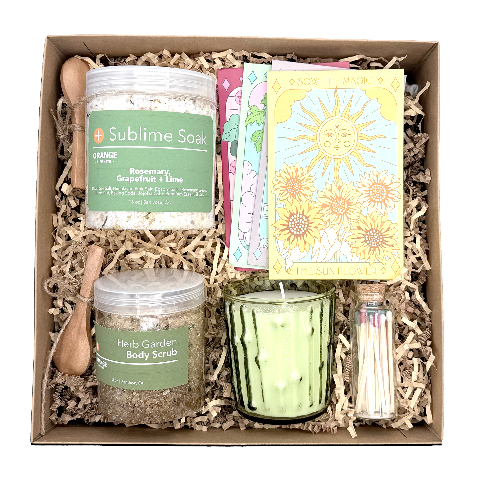 Gift box containing a green Herbal Garden Body Scrub jar, a jar of Sublime Soak bath salts, and a yellow card with a sun and flower illustration.