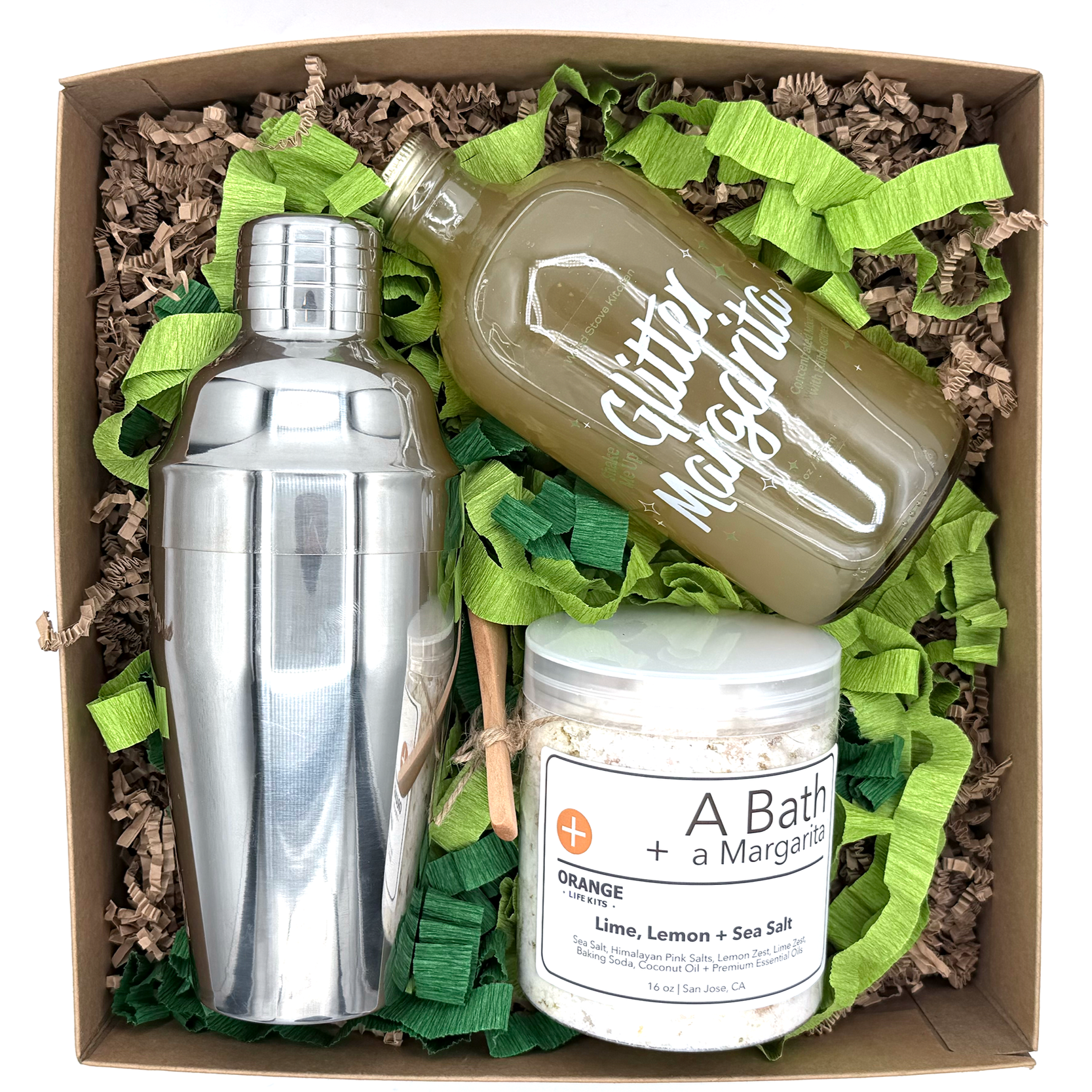 Gift box containing a stainless steel bottle, a glass bottle labeled "Garage" with a beverage, and a jar labeled "A Bath + Margarita" with ingredients for a lime, lemon, and sea salt bath, surrounded by green and brown decorative paper fill.