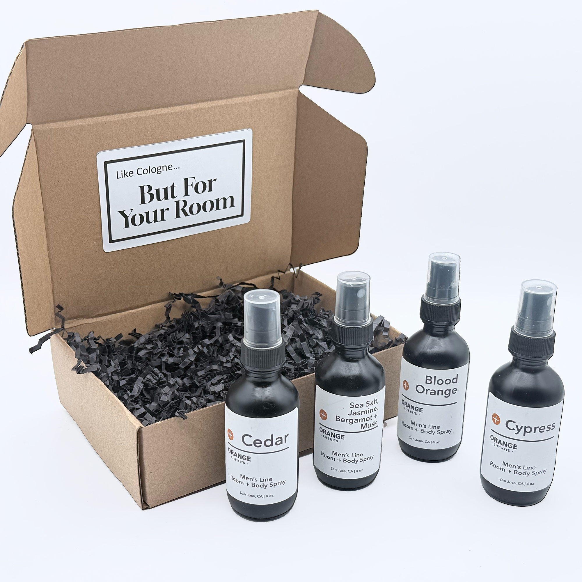 Open cardboard box filled with black shredded paper, containing three small spray bottles with labels for men's room and body spray scents, positioned in front of the box.