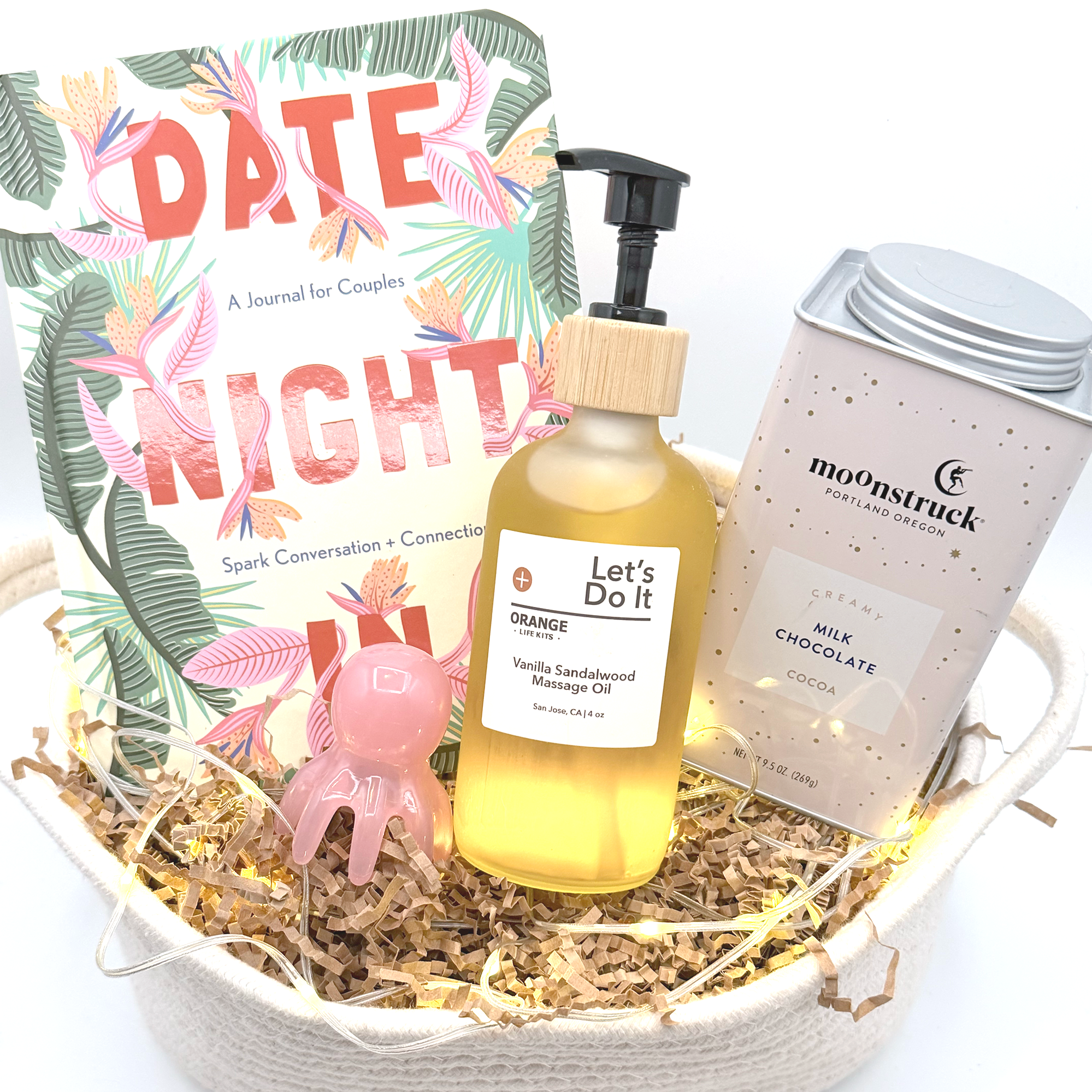Bouquet with a book titled 'Date Night' by Heart to Heart Connection, a bottle of massage oil labeled 'Let's Do It,' a box of Milk Chocolate cookies, and a small pink bear-shaped ornament, all arranged on shredded paper inside a white basket.