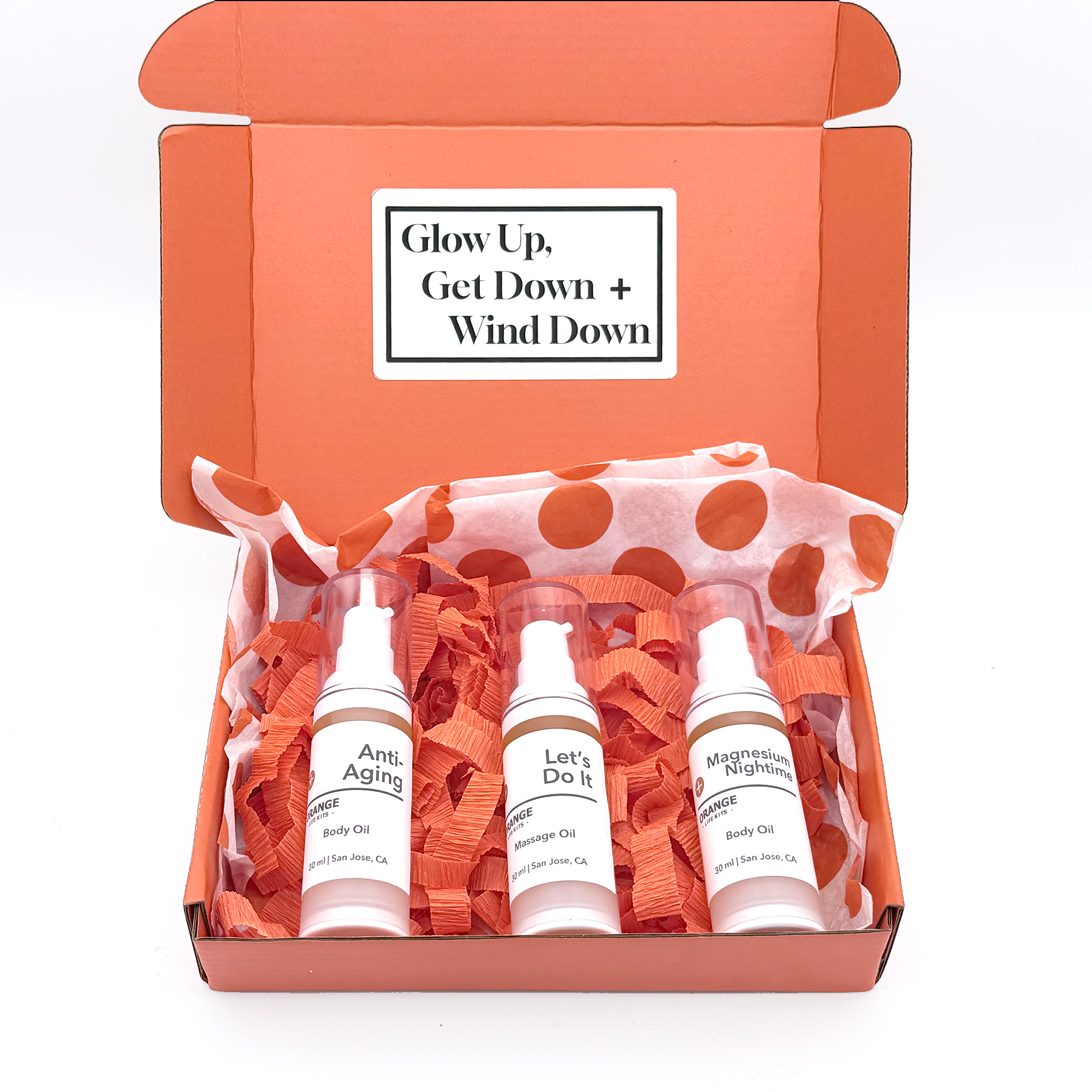 An open orange box with three white bottles labeled 'Anti-Aging', 'Let's Do It', and 'Magnesium Nighttime', containing body oils, placed on orange shredded paper with a polka dot pattern.