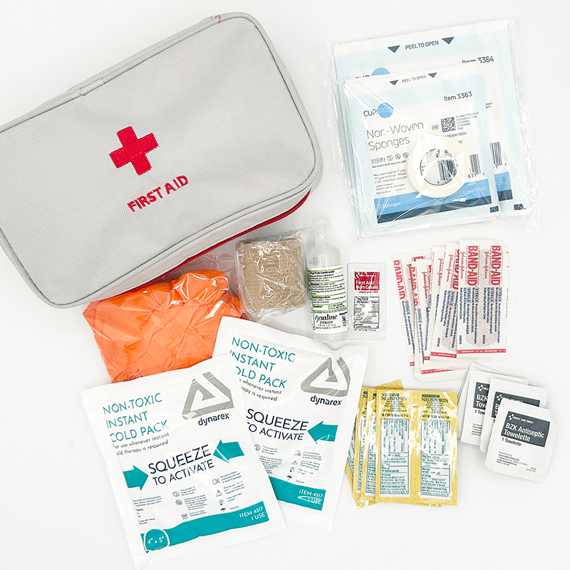 A first aid kit with various medical supplies including antiseptic wipes, bandages, alcohol wipes, and non-woven sponges, arranged on a white surface.