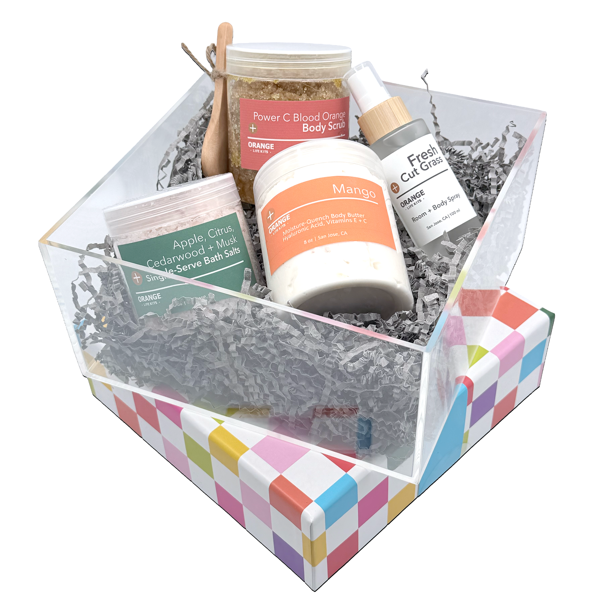 A gift box filled with various health and wellness products including a sugar scrub, body spray, bath salts, and body butter, arranged on crinkle paper with colorful diamond patterns on the box.