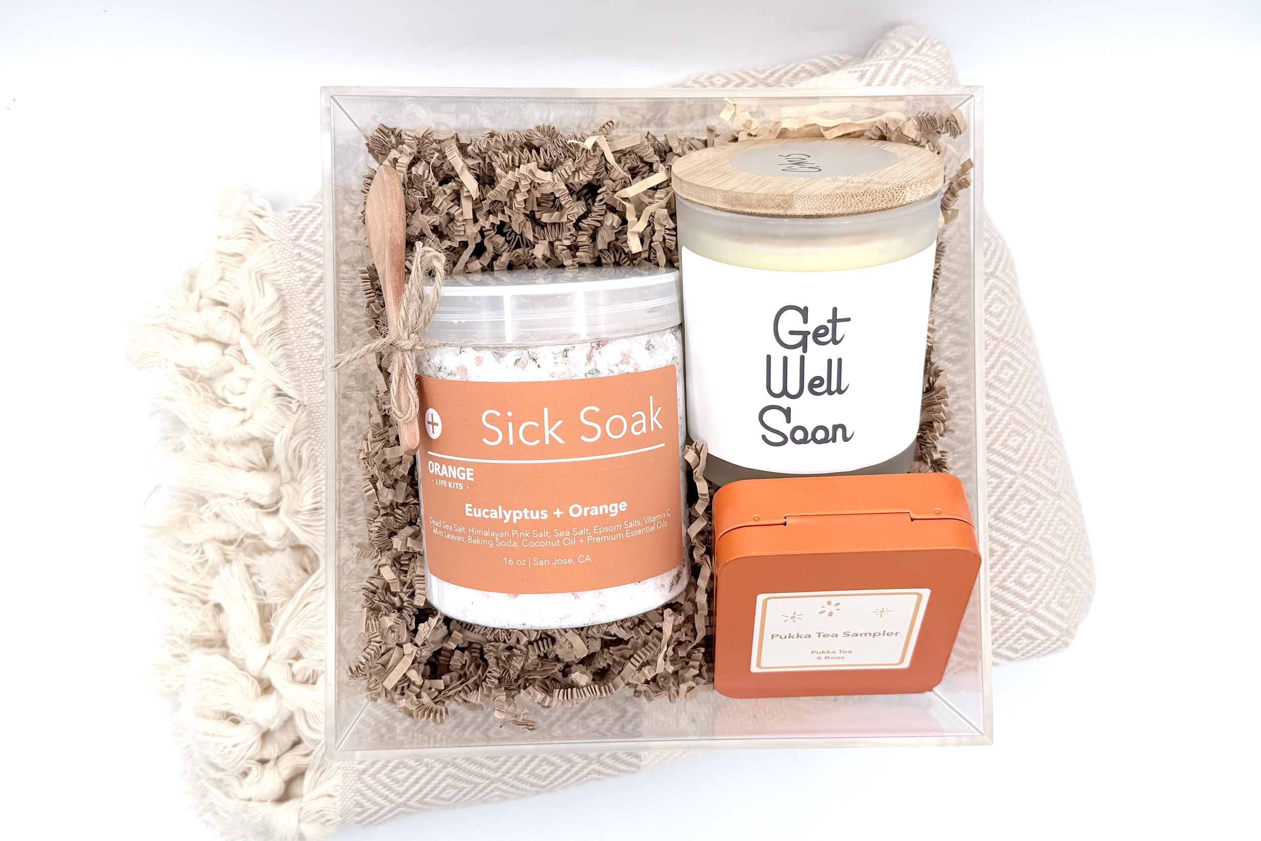 Gift set including a candle labeled 'Get Well Soon', a jar of bath salts labeled 'Sick Soak', and a pumka tea sampler, all arranged on a decorative surface with shredded paper filler.