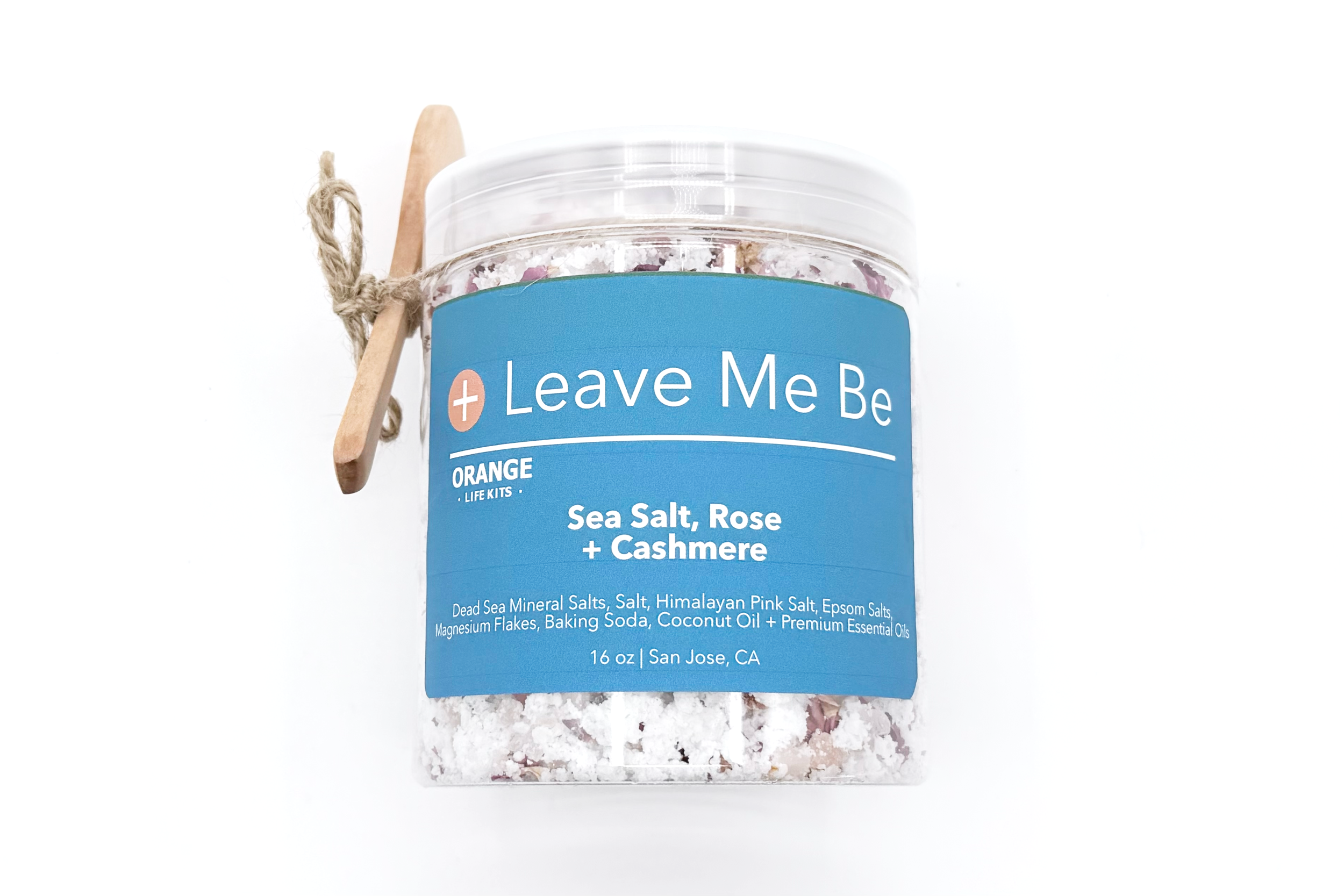 A clear plastic jar of Leave Me Be sea salt, rose, and cashmere bath salts with a blue label and a wooden tongue depressor tied with string attached to the jar.