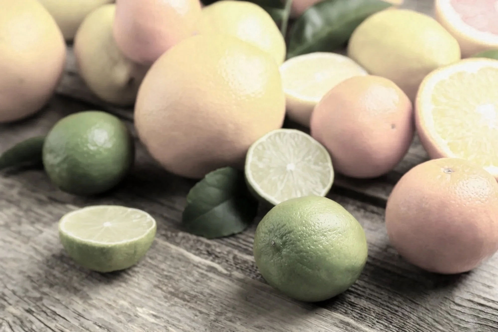 A variety of fresh citrus fruits including whole limes, lemons, and halved lemons, displayed on a wooden surface with some green leaves in the background.