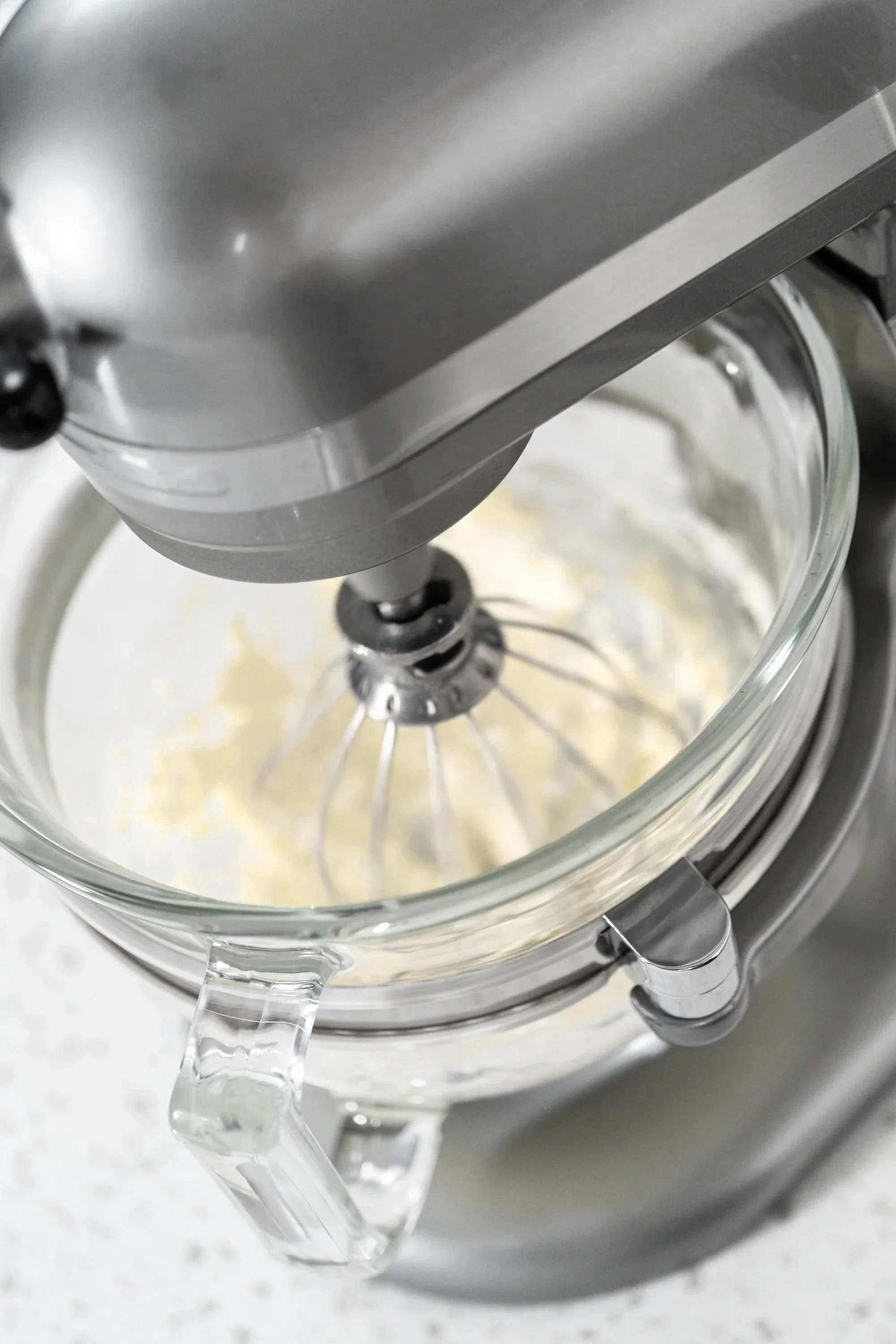 Close-up of a stand mixer with glass bowl, mixing egg whites or cream, on a kitchen counter.