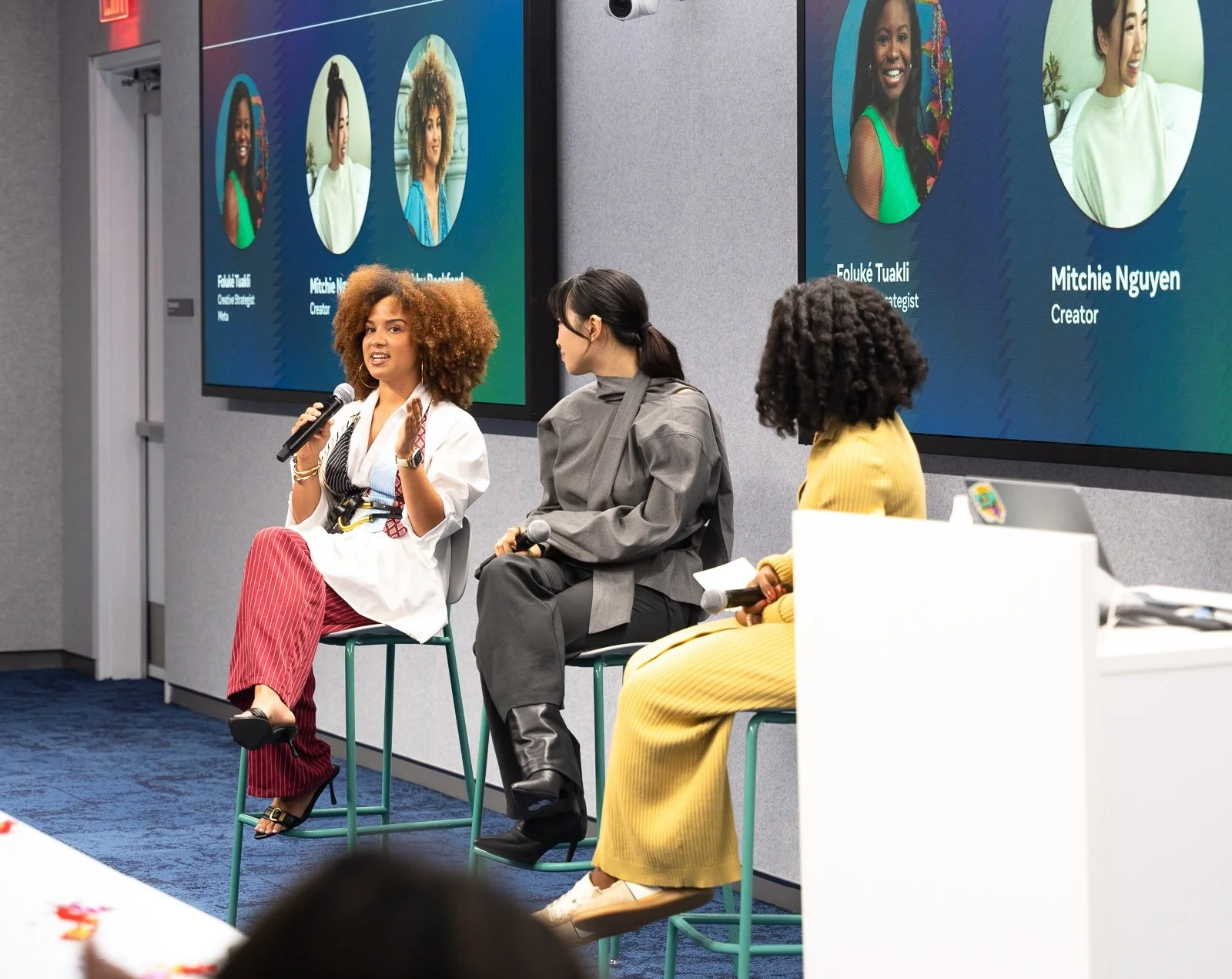 Three women are seated on stage during a panel discussion, each holding a microphone, with large screens behind them displaying their names and titles. The woman on the left, with curly hair, is speaking, while the other two listen. The screens displ
