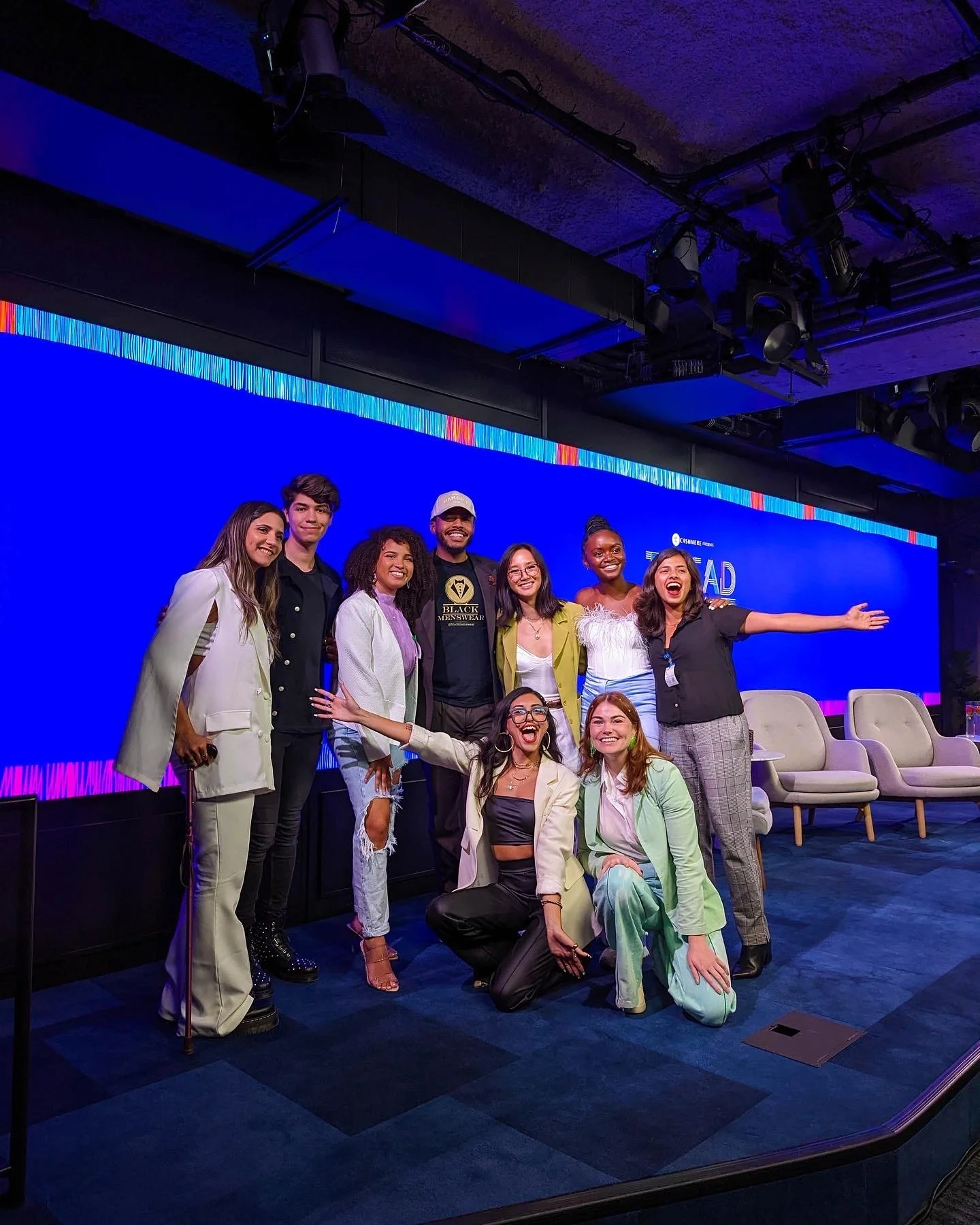 Group of nine diverse people smiling and posing together on a stage with a blue digital background, some standing and some kneeling, in a professional or conference setting.