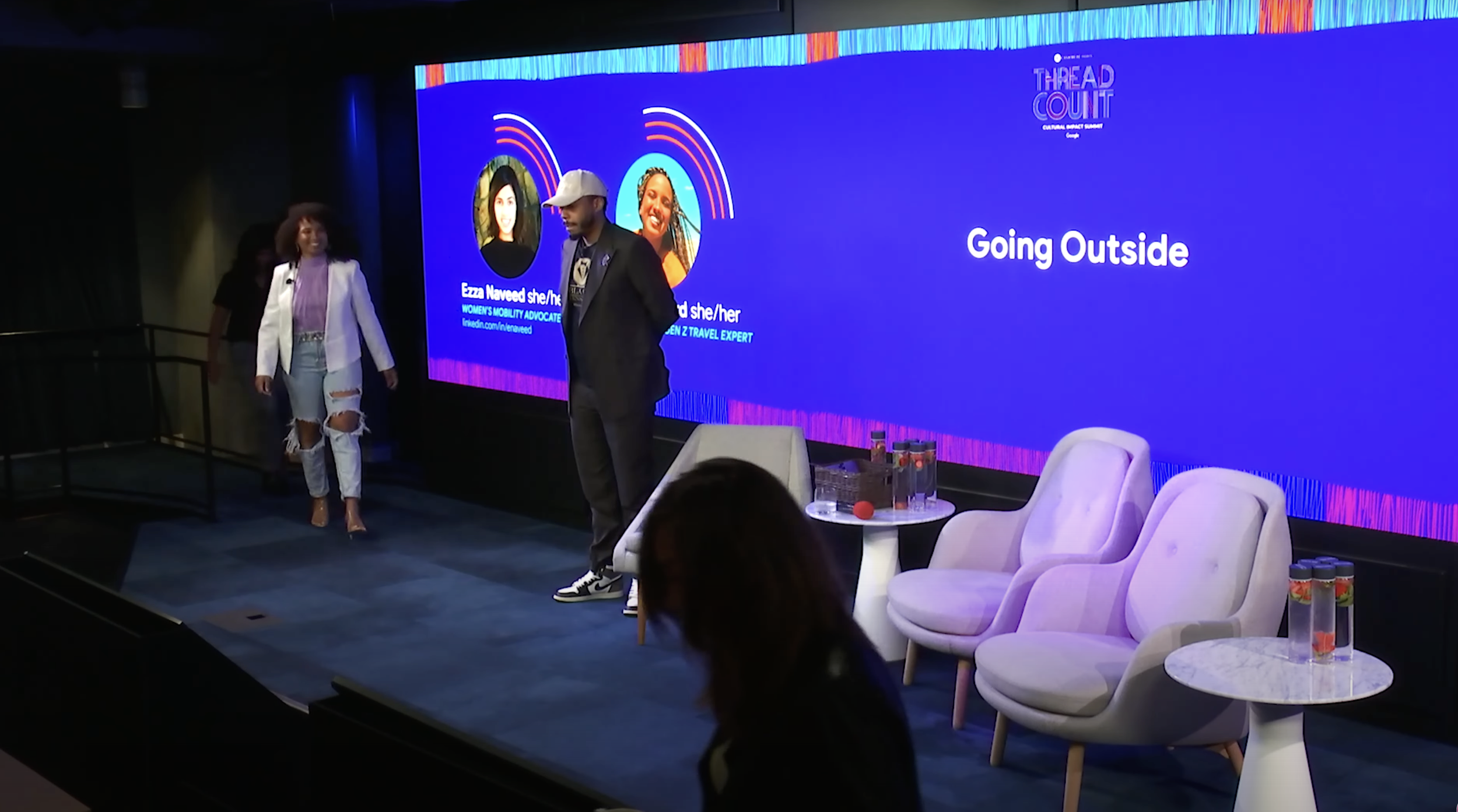A stage with three white chairs, a small table with water bottles, and a large screen displaying the topic "Going Outside" during a conference or panel discussion.