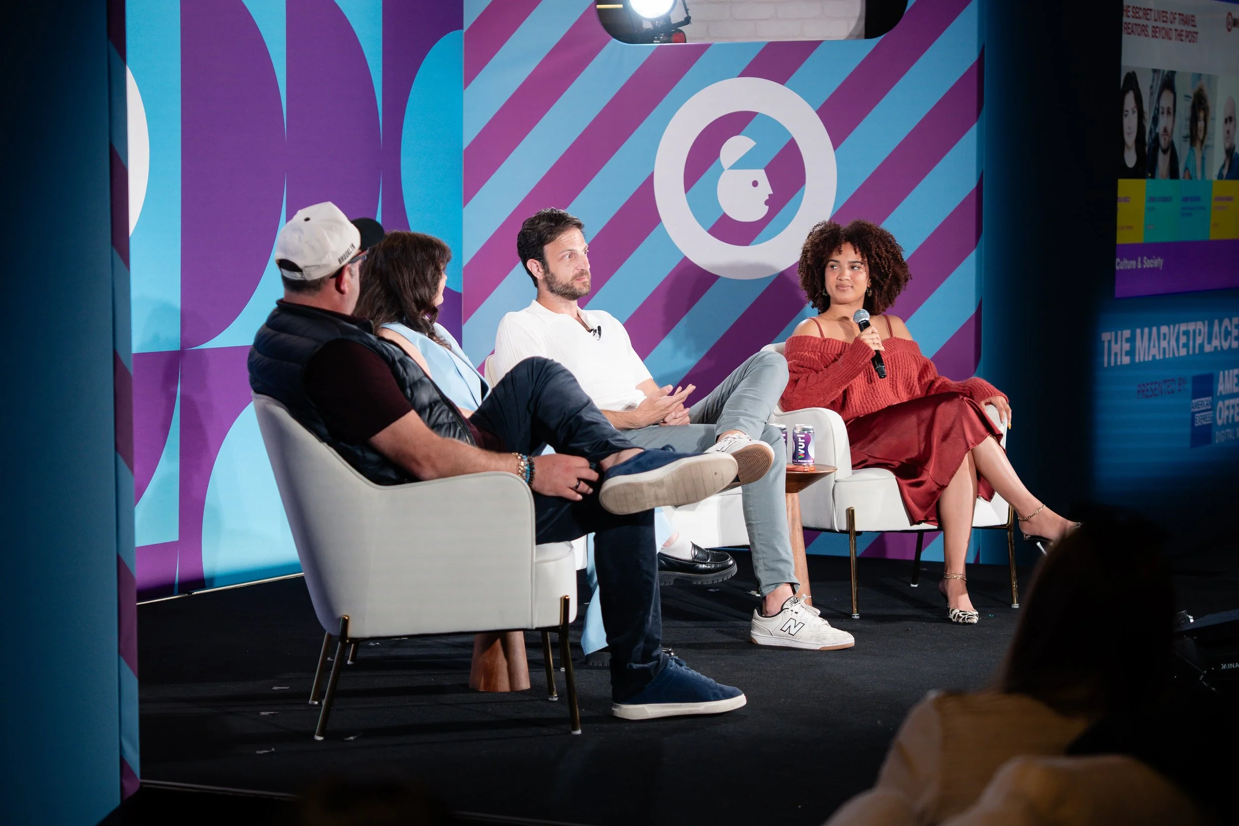 Four people sitting on stage in a panel discussion, with a purple and blue geometric background and a logo of a stylized face in profile.