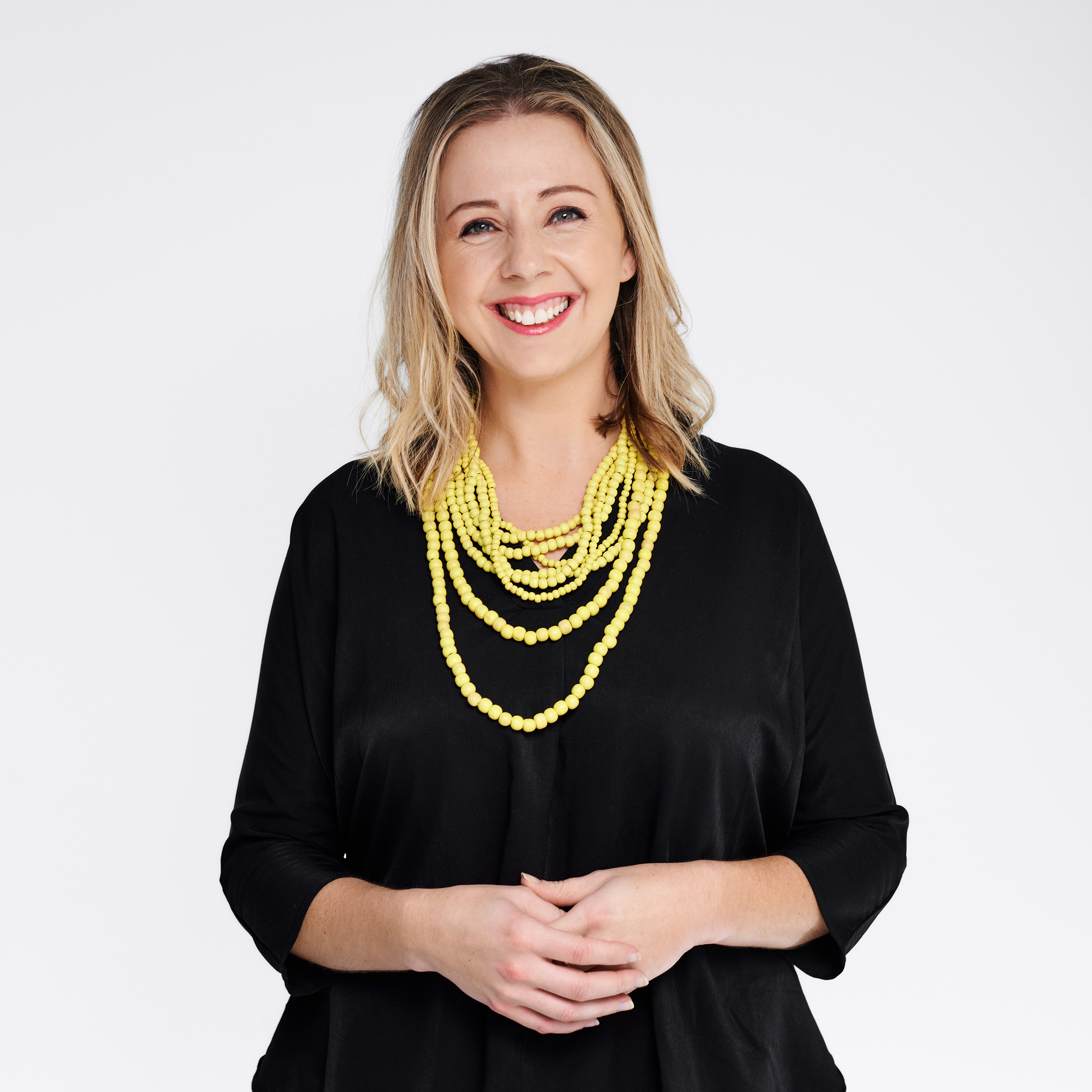 A smiling woman with blonde hair wearing a black top and a multi-strand yellow bead necklace.