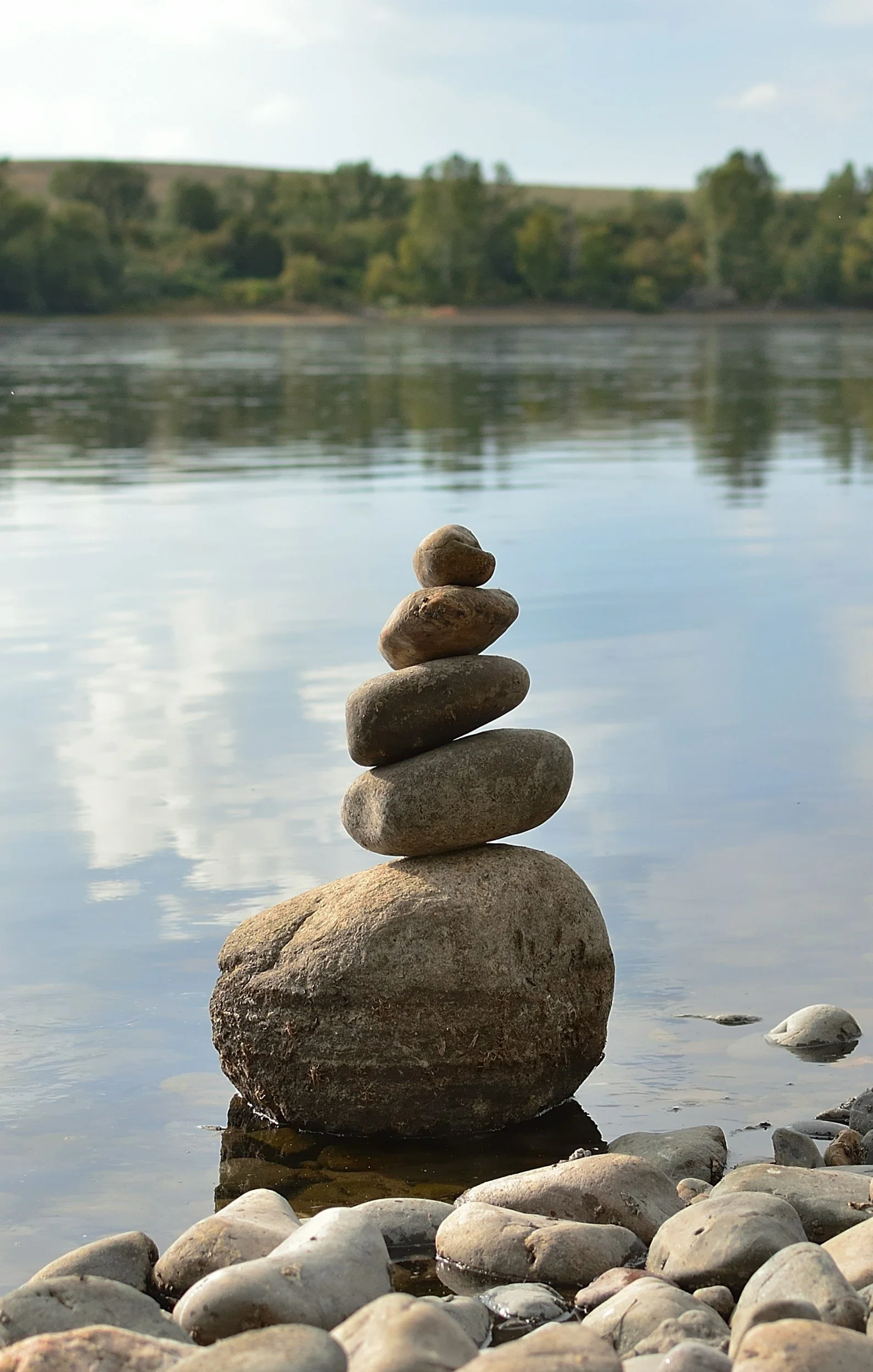 A tall stack of five rocks balanced on a large baseline rock near a calm river, with a background of trees, fields, and a partly cloudy sky.