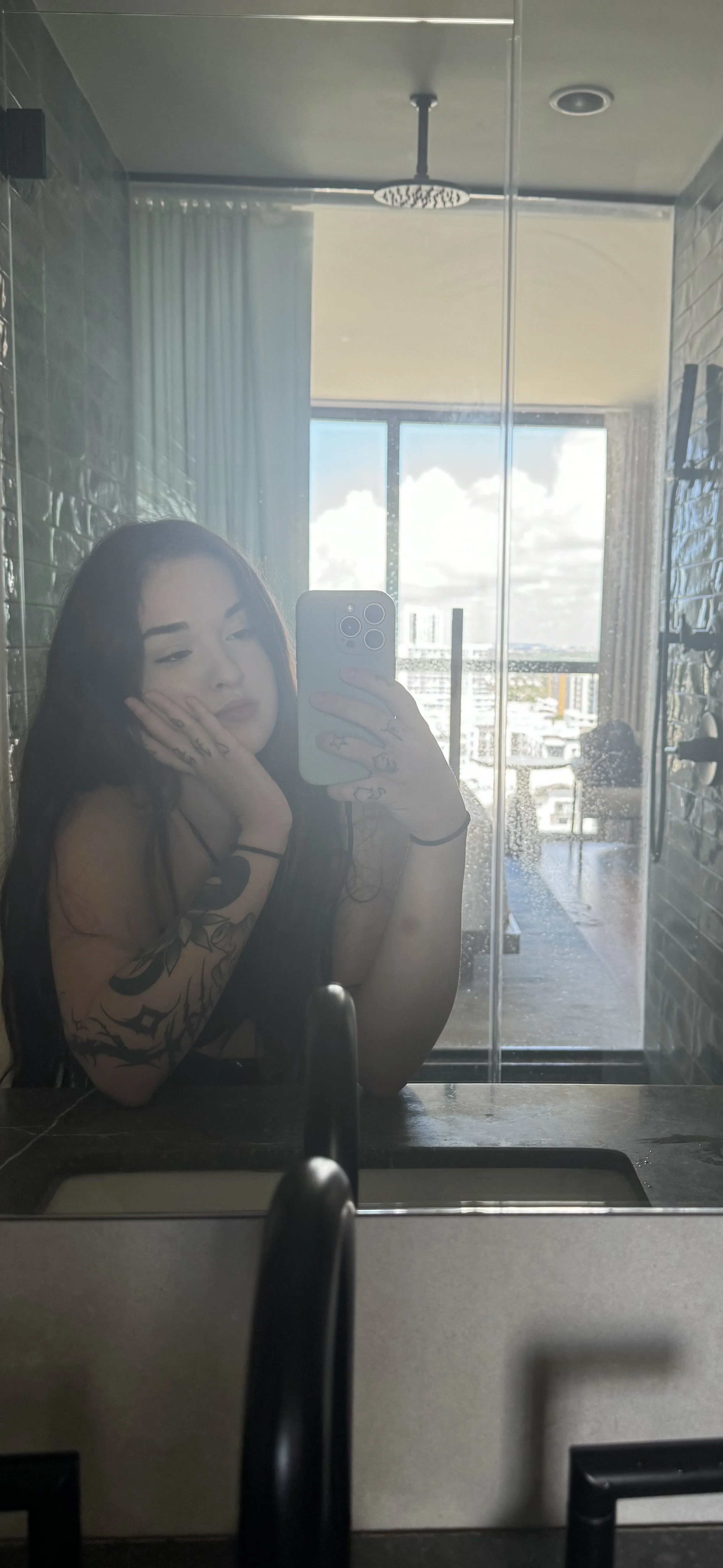 Woman with long dark hair and tattoos on her arm taking a mirror selfie in a bathroom with a city view, cloudy sky outside.