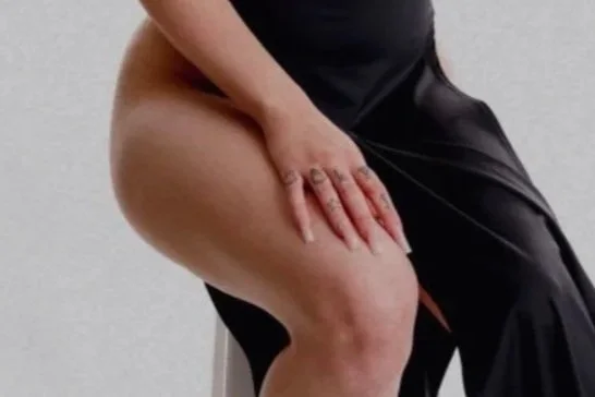 Close-up of a person sitting, wearing black clothing, with one hand on their thigh, against a plain gray background.