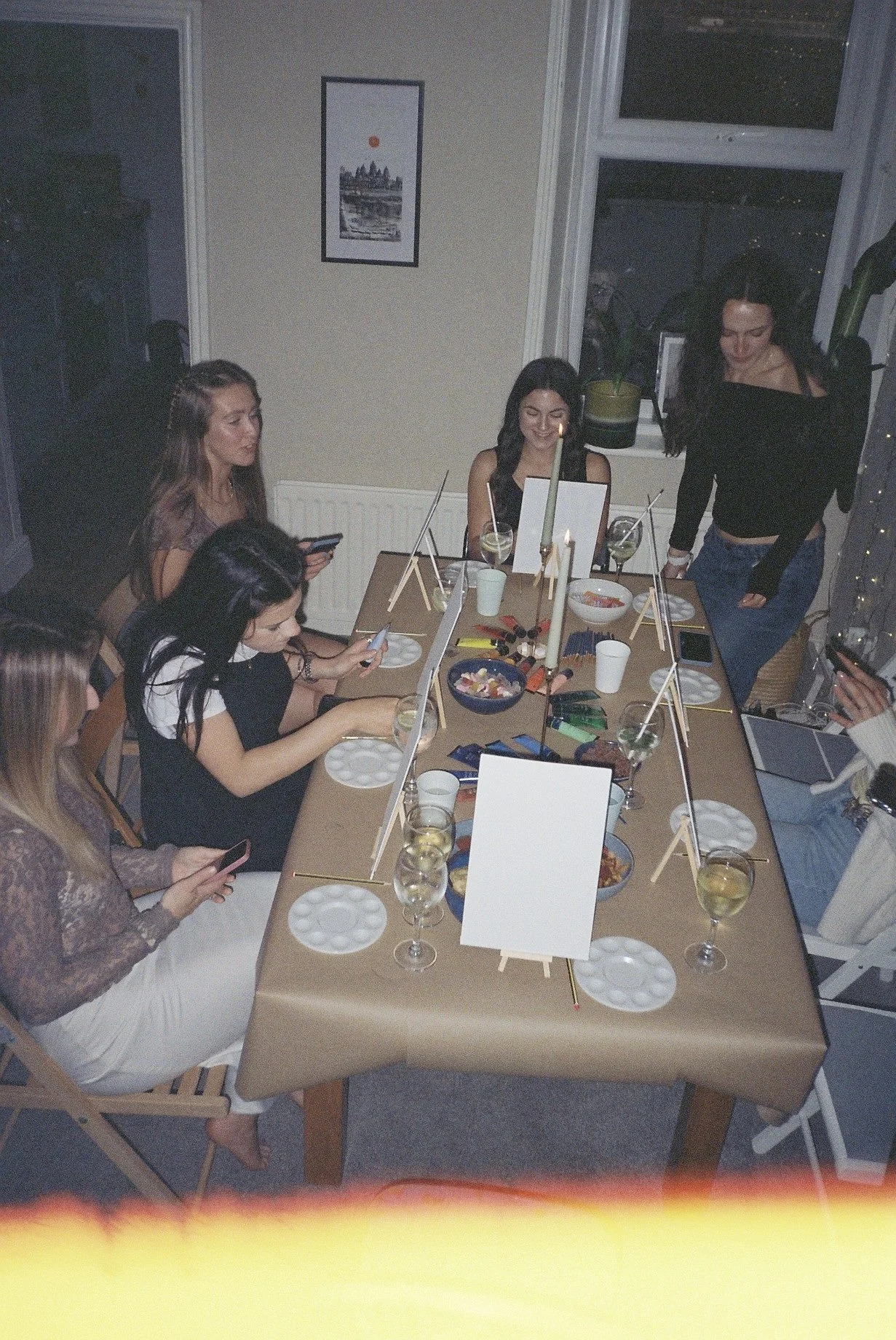 A young woman with long dark hair sitting on the floor against a yellow-green background, hugging her knees with her arms, wearing a bracelet, socks, and white lace-up boots.