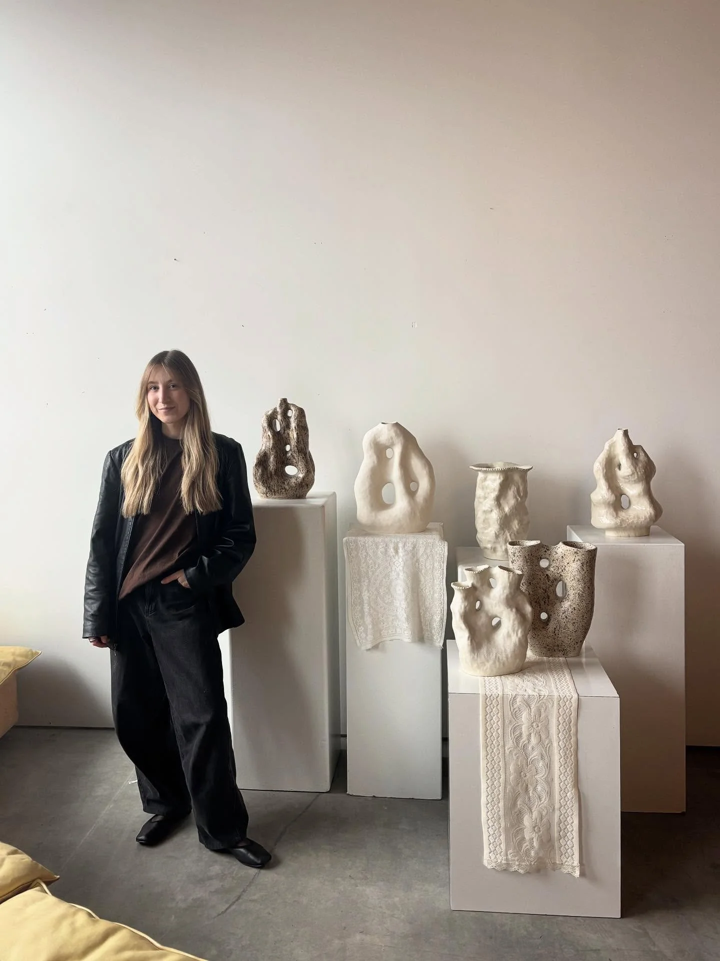 Someone Ceramics.  A ceramic artist in Victoria, A woman with long blonde hair, wearing a black jacket and dark pants, stands next to white pedestals displaying ceramic sculptures with holes and textures, with some textiles draped over the pedestals.