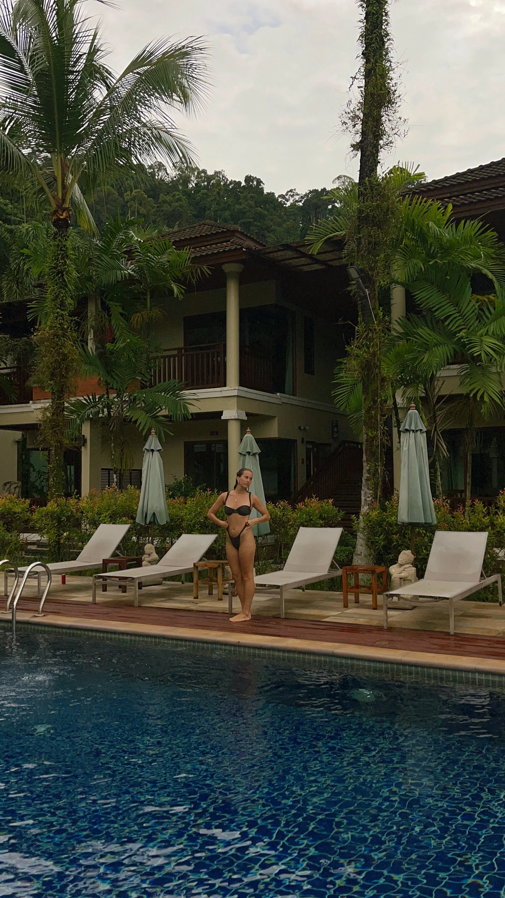 Woman in black swimsuit standing poolside with lounge chairs and umbrellas, lush greenery and a two-story house in the background.