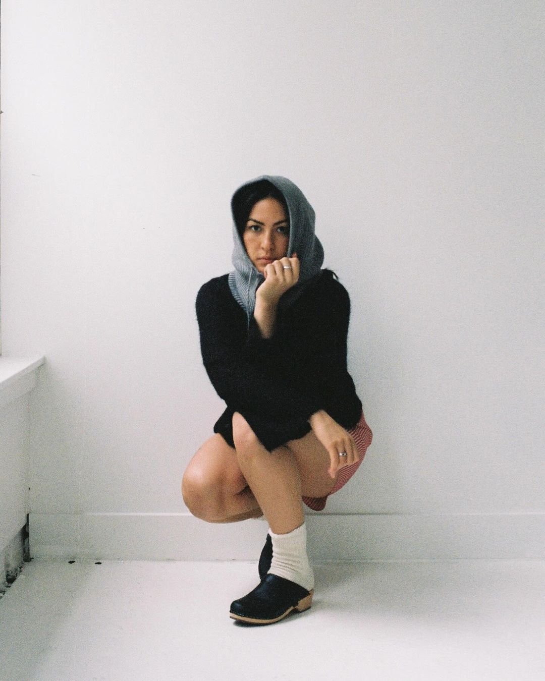 A woman with dark hair, wearing a grey hoodie, black sweater, red checkered shorts, and black shoes with beige socks, squatting in front of a plain white wall, gazing at the camera.