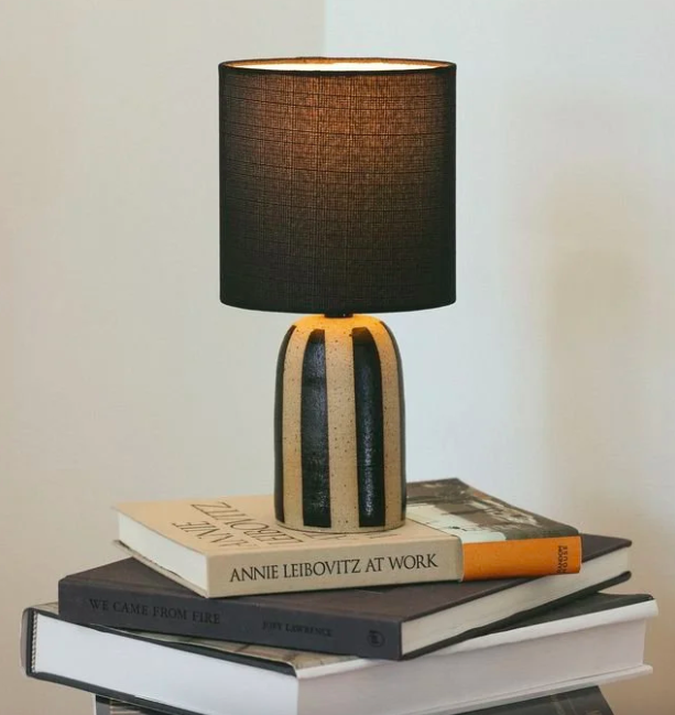 Natalya Jade Ceramics,  a ceramic artist in Victoria, BC. making ceramic lamps. A table lamp with a black lampshade and a textured beige base with dark vertical stripes, placed on top of a small stack of books and magazines on a black surface.