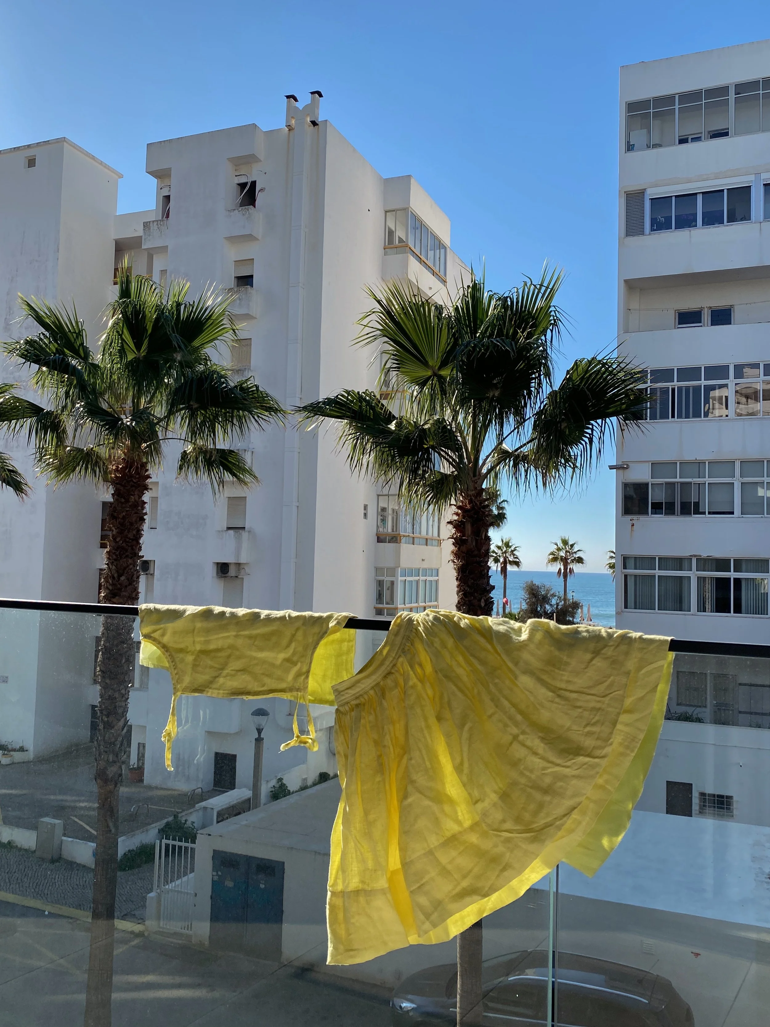Shelter Clothing, linen sets made in Canada. Yellow clothing hanging on a clothesline outside a window, with white apartment buildings and palm trees in the background under a blue sky.