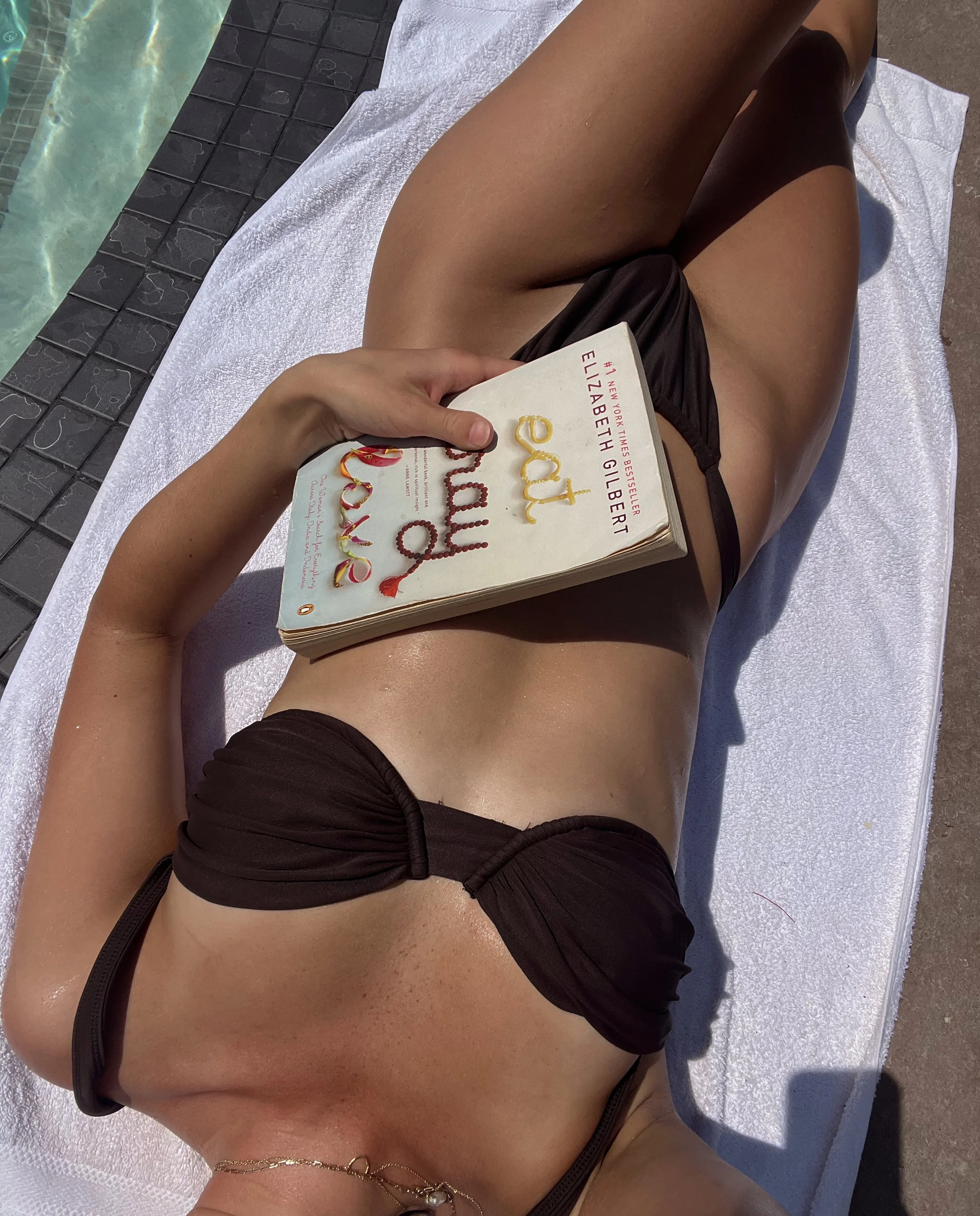 A woman relaxing by the poolside, lying on a white towel. She is wearing a black bikini and is holding an open book titled 'Eat Joyfully' by Elizabeth Gilbert.