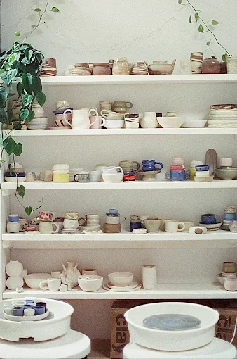 Caitlin Prince Ceramics. A ceramic artist in Victoria, BC. White shelves with various stacked and arranged pottery bowls, cups, and plates, with a green plant on the left side.