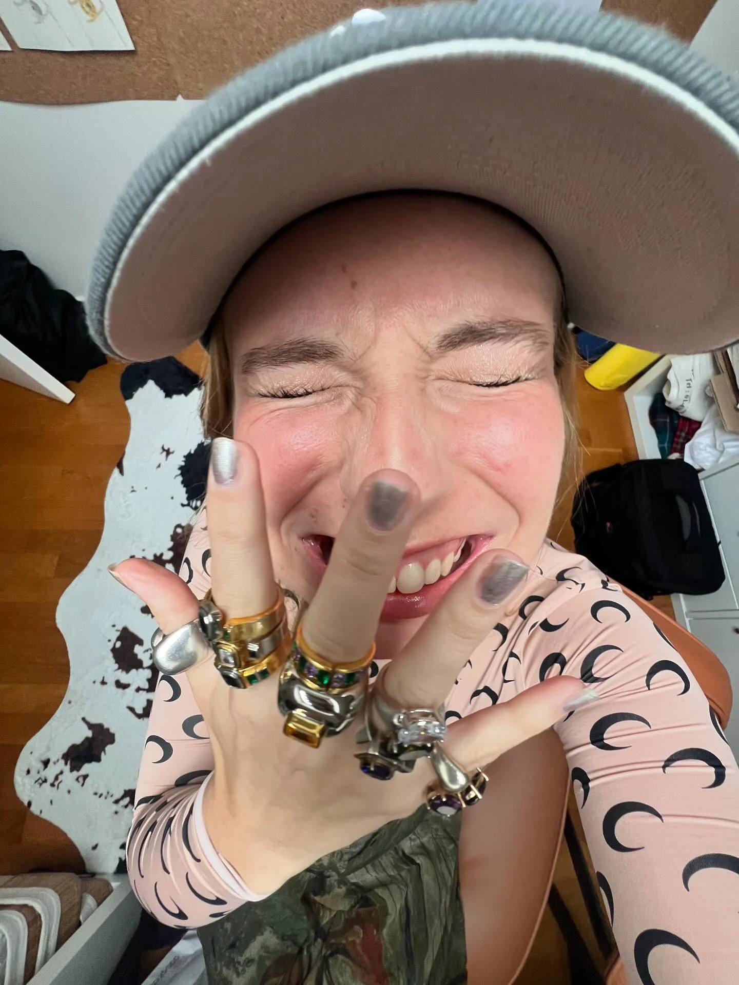 A woman taking a selfie from above, smiling with her eyes closed, wearing a large hat, multiple rings on her fingers, and a patterned pink top. The background shows a room with furniture and a cow print rug.