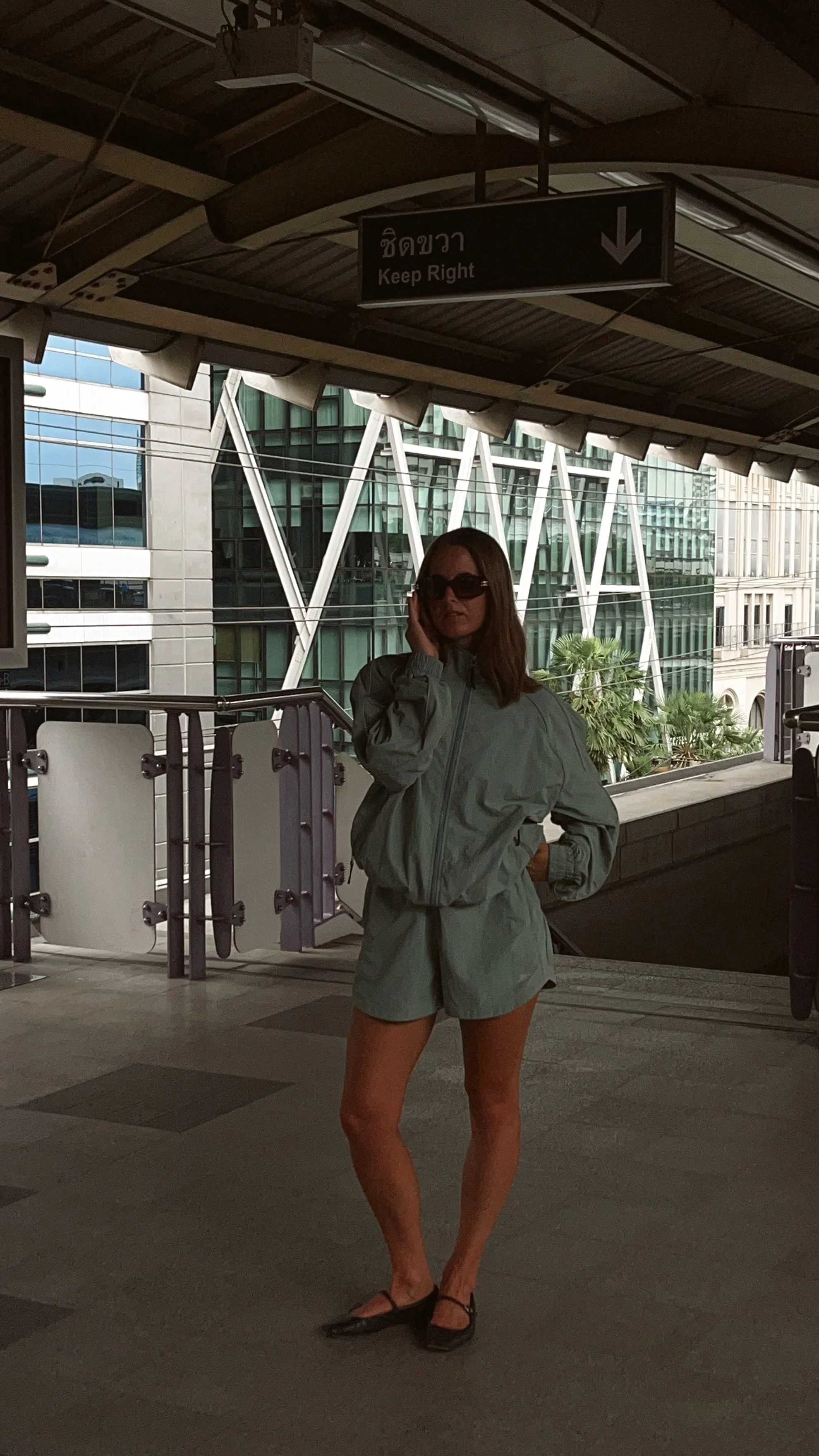 Planet Nusa. A woman wearing sunglasses, a mint green jacket, and green shorts standing on an outdoor elevated platform with buildings and tropical plants in the background. She is looking away from the camera and holding her hand up to her face.