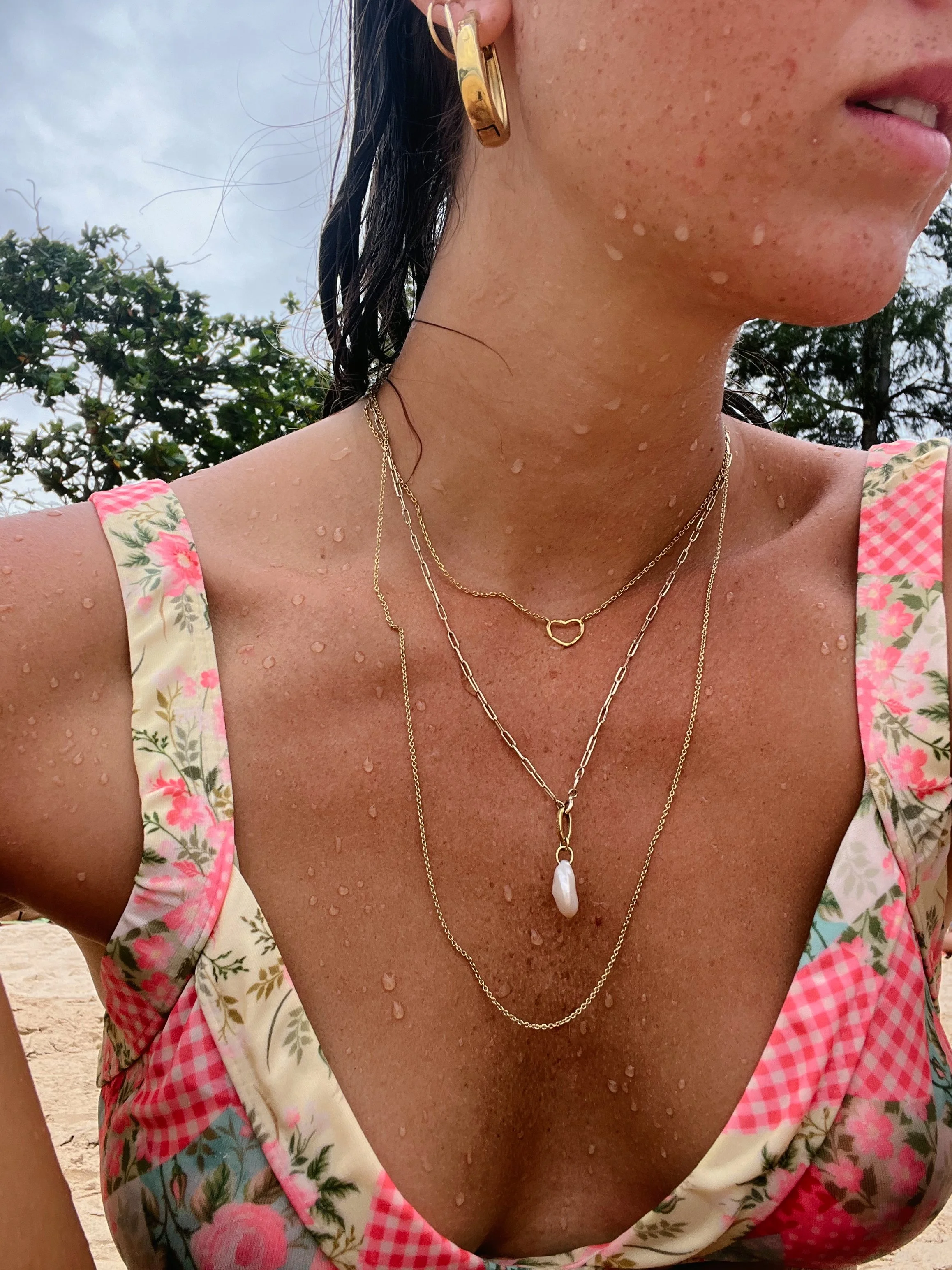 Close-up of a woman's bare shoulder and neck with layered gold necklaces, including a heart-shaped pendant and a pearl pendant, wearing gold hoop earrings and a floral sleeveless top, water droplets on skin, outdoors with trees and cloudy sky in back
