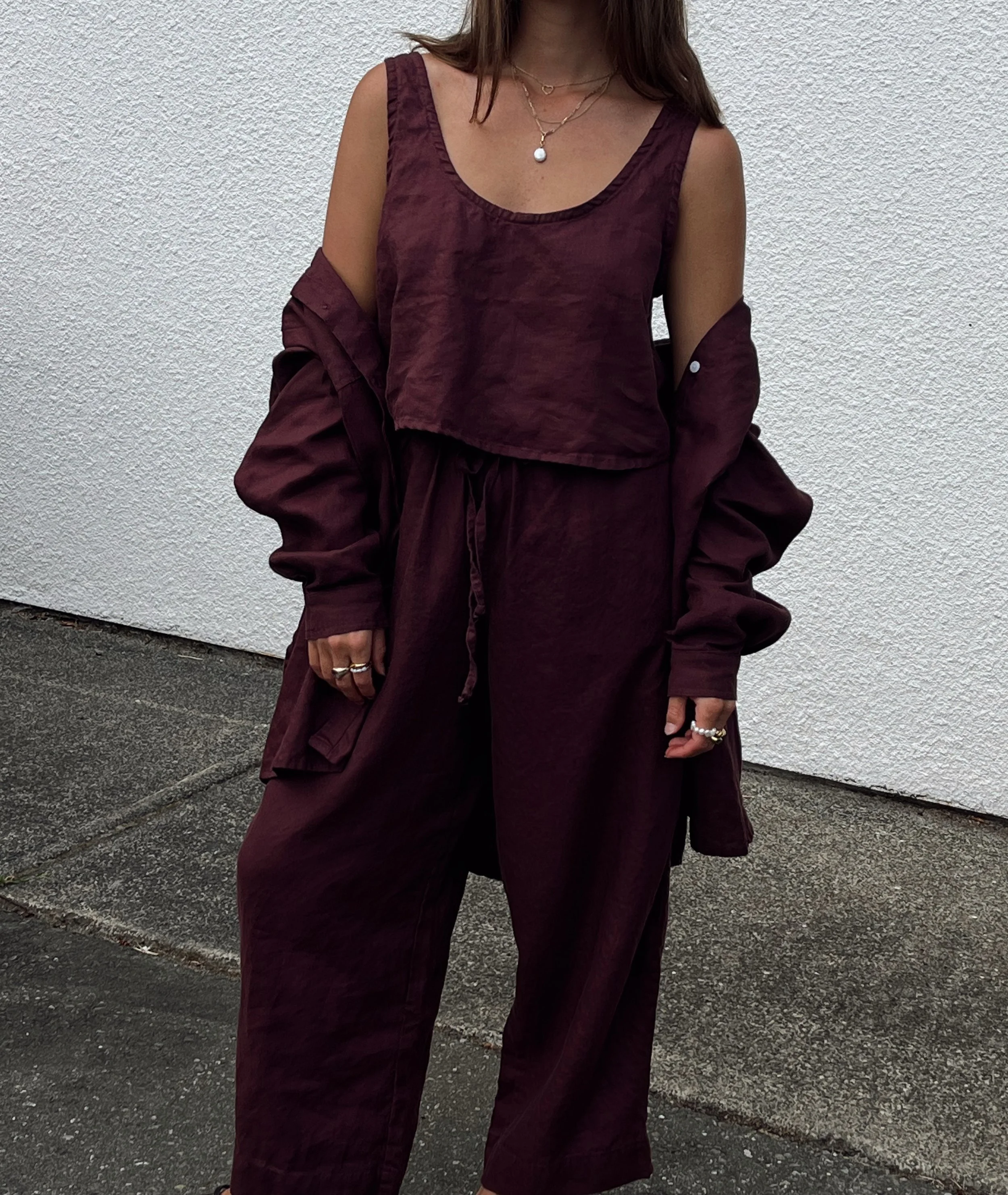 Shelter Clothing, linen sets made in Canada. Woman wearing a matching burgundy sleeveless top, loose pants, and a jacket draped over her shoulders, standing against a white textured wall.