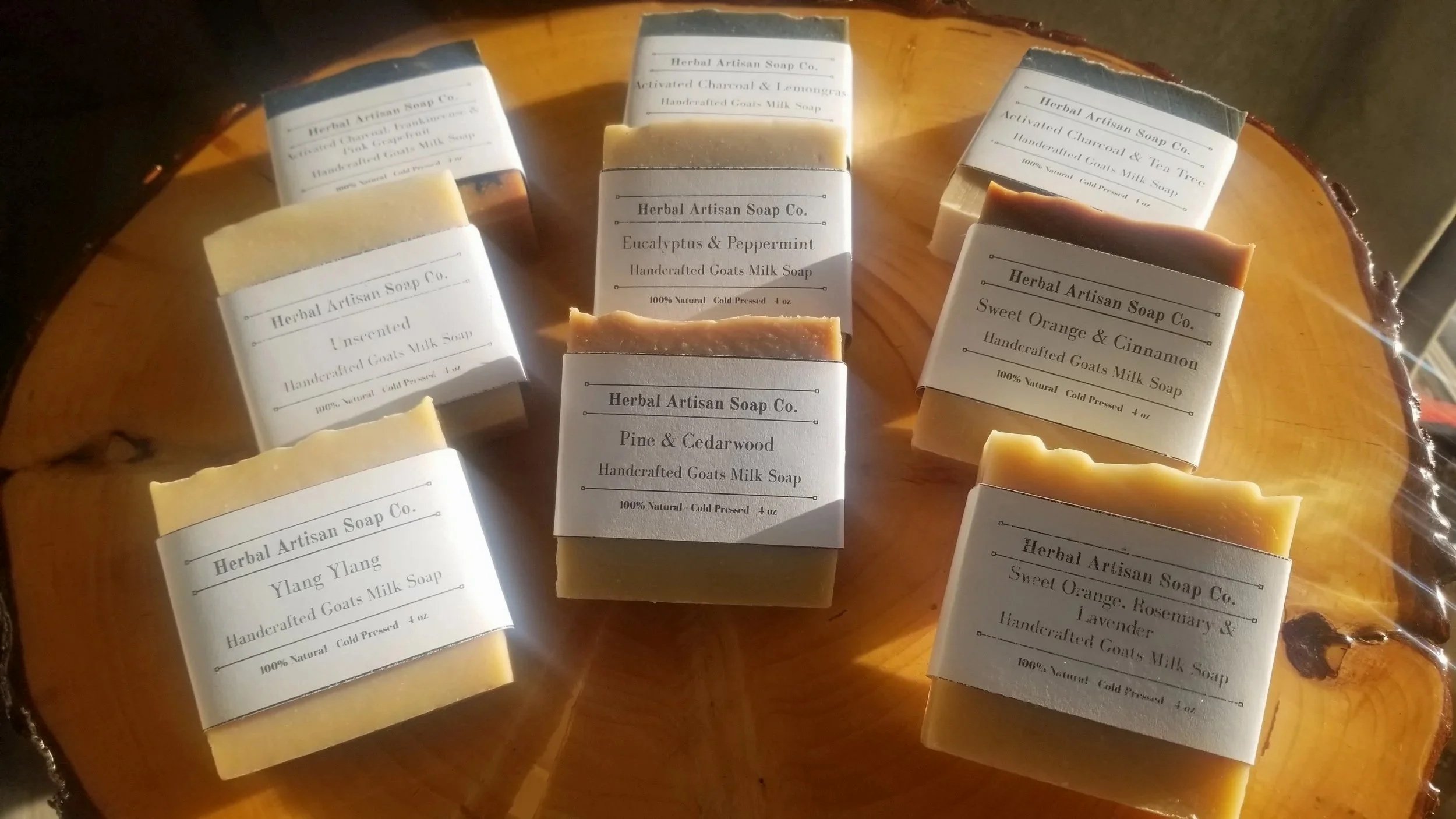 Several handcrafted soap bars with white labels on a wooden surface. The soaps are in different colors and scents, including eucalyptus & peppermint, sweet orange & cinnamon, pine & cedarwood, Ylang Ylang, and a scented goat milk soap. The labels indicate they are natural, cold pressed, and made by Herbal Artisan Soap Co.