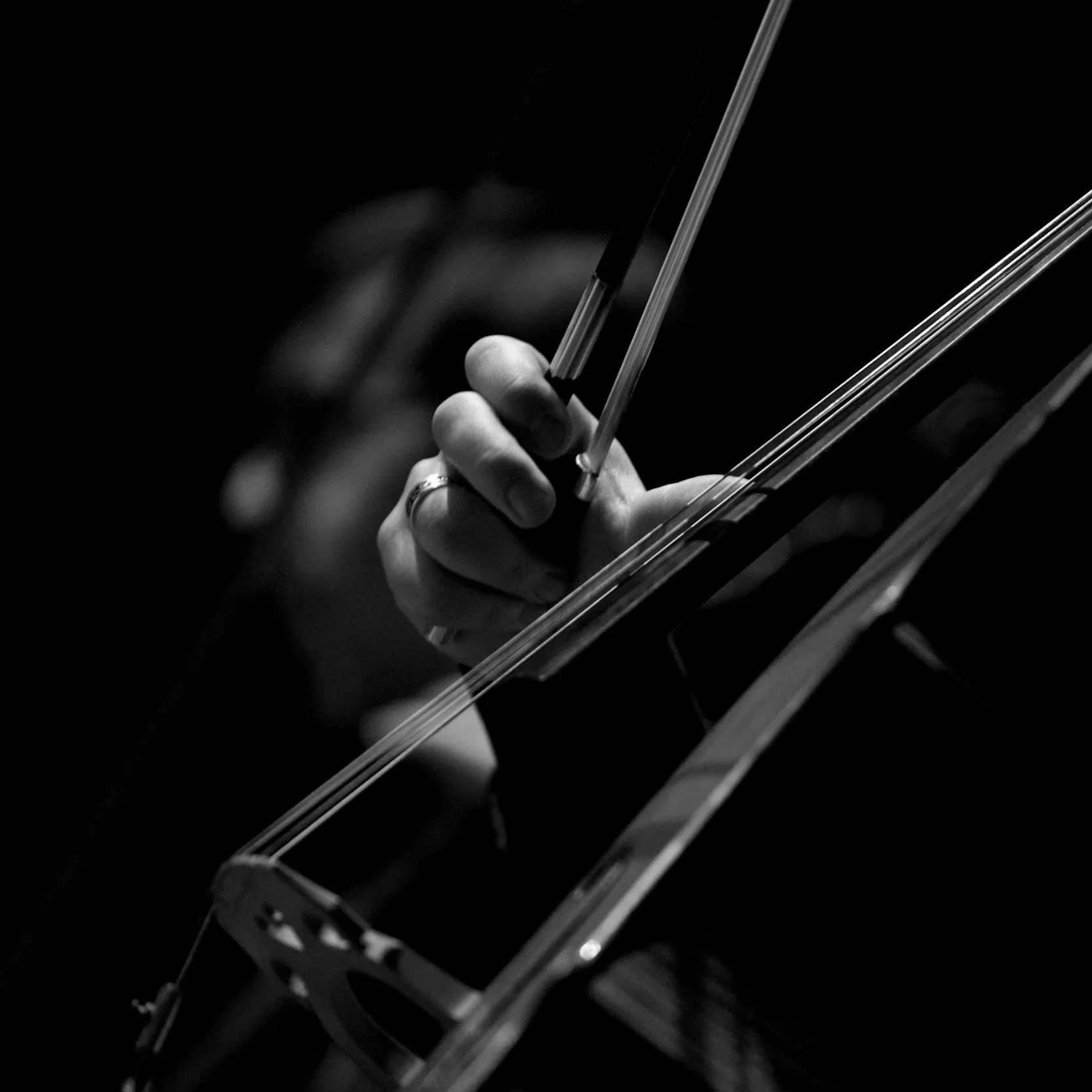 Close-up of a hand holding a violin bow, playing on a violin, in black and white with a dark background.