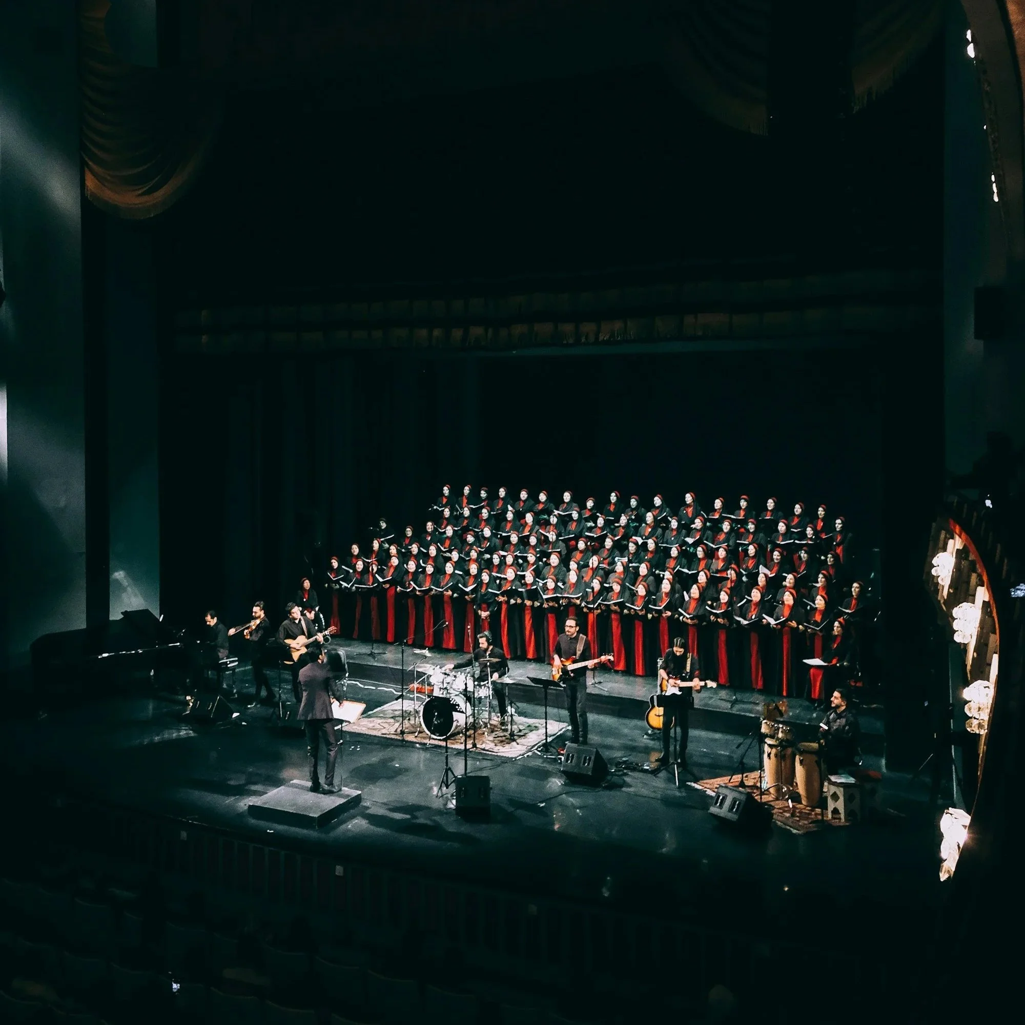 A choir dressed in black robes with red accents, standing on risers onstage in a theater. Musicians are performing in front of the choir, including a conductor, guitarists, a drummer, and other players. The stage is lit, with a dark backdrop and ornate side décor.