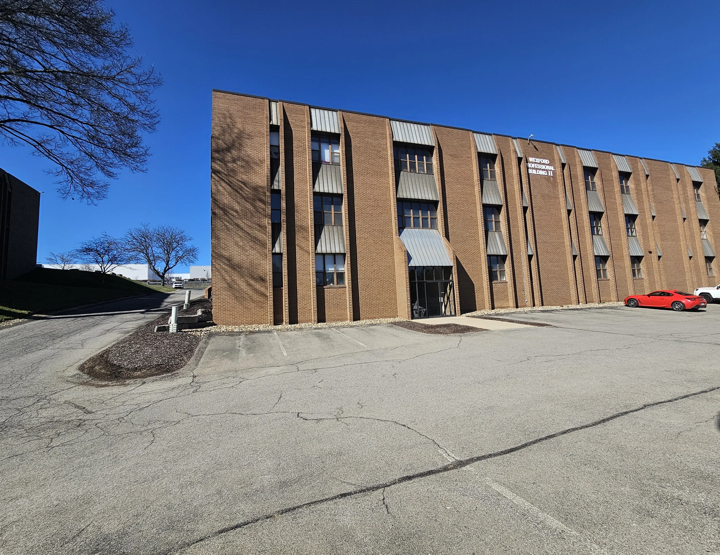 Brick building with metal awnings, parking lot with a red sports car and a white vehicle, leafless trees, clear blue sky.