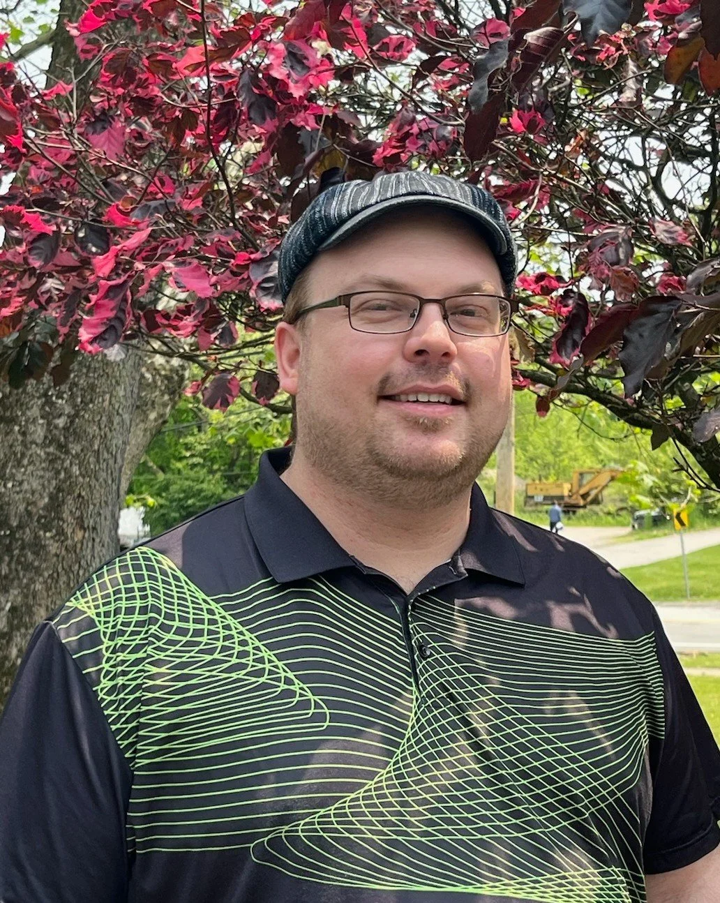 A man wearing glasses, a black and neon green patterned polo shirt, and a striped cap outdoors, standing in front of a tree with red and dark purple leaves.