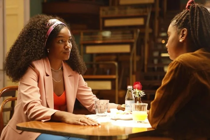 Cena do filme Um ano Inesquecível - Outono. Na imagem temos a figura de Iza, mulher negra, de cabelho cacheado longo, vestindo uma roupa rosa. Ao seu lado está Anna Júlia (Gabz), uma jovem negra, com locs braids. Elas estão se olhando.