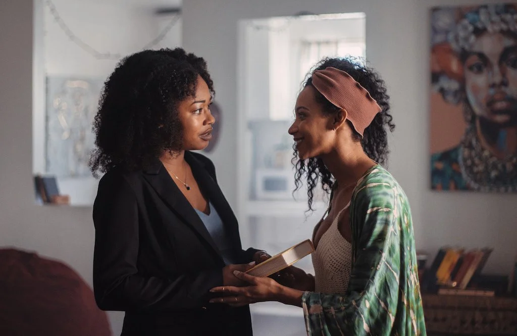 Cena do filme Tender. Na esquerda temos Kiara (Farelle Walker), uma mulher negra de cabelo cacheado. Ao seu lado está Lulu (Trishauna Clarke), outra mulher negra de cabelo cacheado. Elas estão se olhando e de mãos dadas.