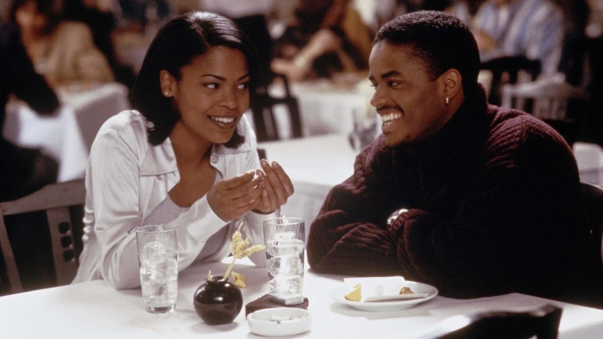 Cena do filme Love Jones. Na imagem temos a imagem de Nina (Nia Long), uma mulher negra, de cabelo curto preto. Ao seu lado está Darius (Larenz Tate), homem negro, cabelo preto raspado. Eles estão em um restaurante, ao redor de várias mesas.