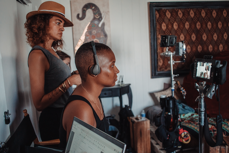 Cena dos bastidores do filme Tender. Na imagem temos Felicia Pride, diretora do curta-metragem, uma mulher negra, cabelo raspado e vestindo uma regata preta. Ao seu lado está sua assistente de gravação, uma mulher negra de cabelo cacheado.